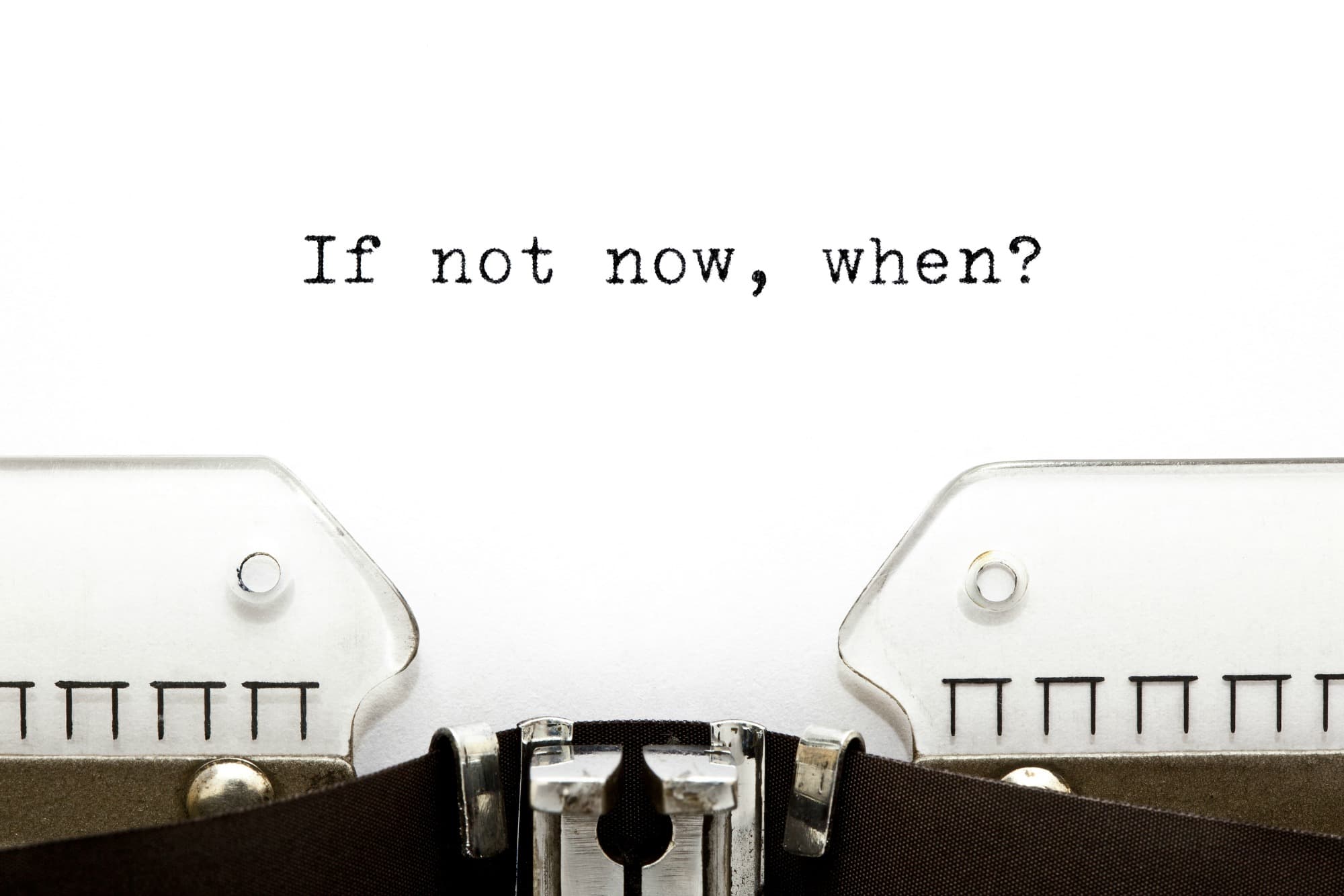 Close-up of a typewriter with a sheet of paper displaying the words If not now, when? typed in black ink at the top of the page.