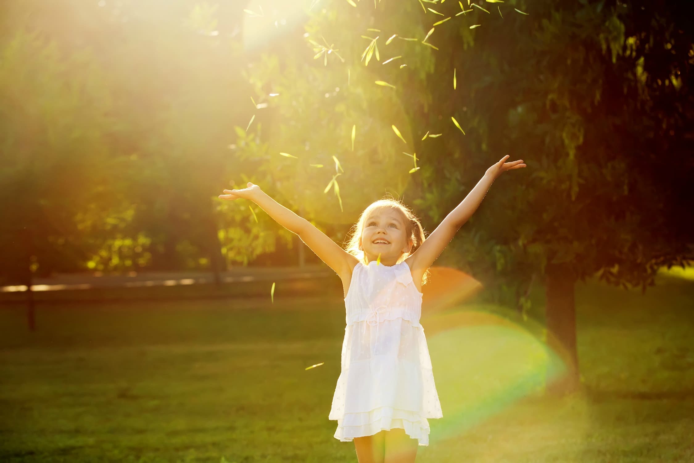 A young girl in a white dress stands on grass with arms raised, smiling joyfully in bright sunlight, surrounded by green trees and illuminated by a warm, golden glow.