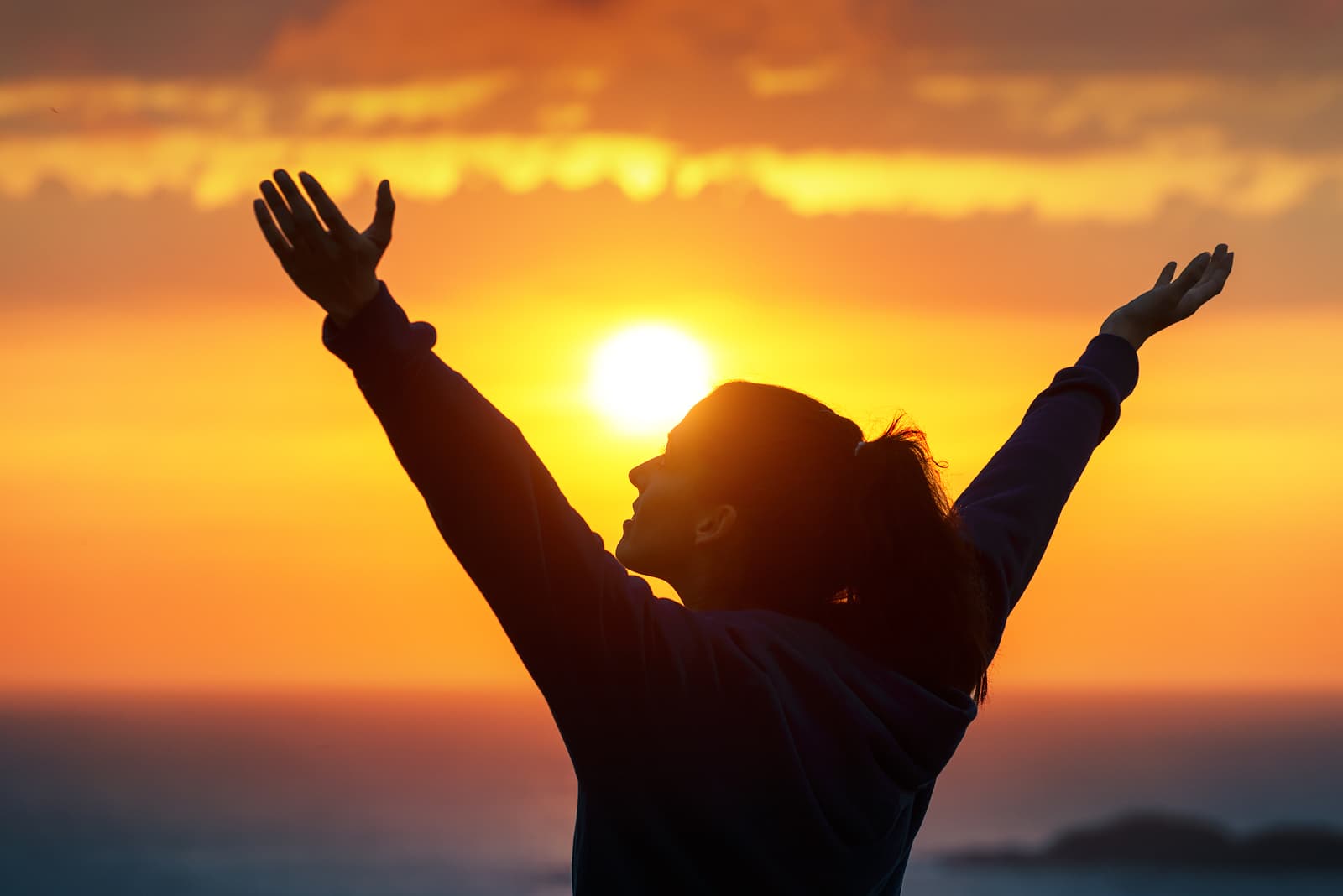 A person stands with arms raised toward the sky, silhouetted against a vibrant orange and yellow sunset over the ocean. The scene conveys a sense of freedom and joy.