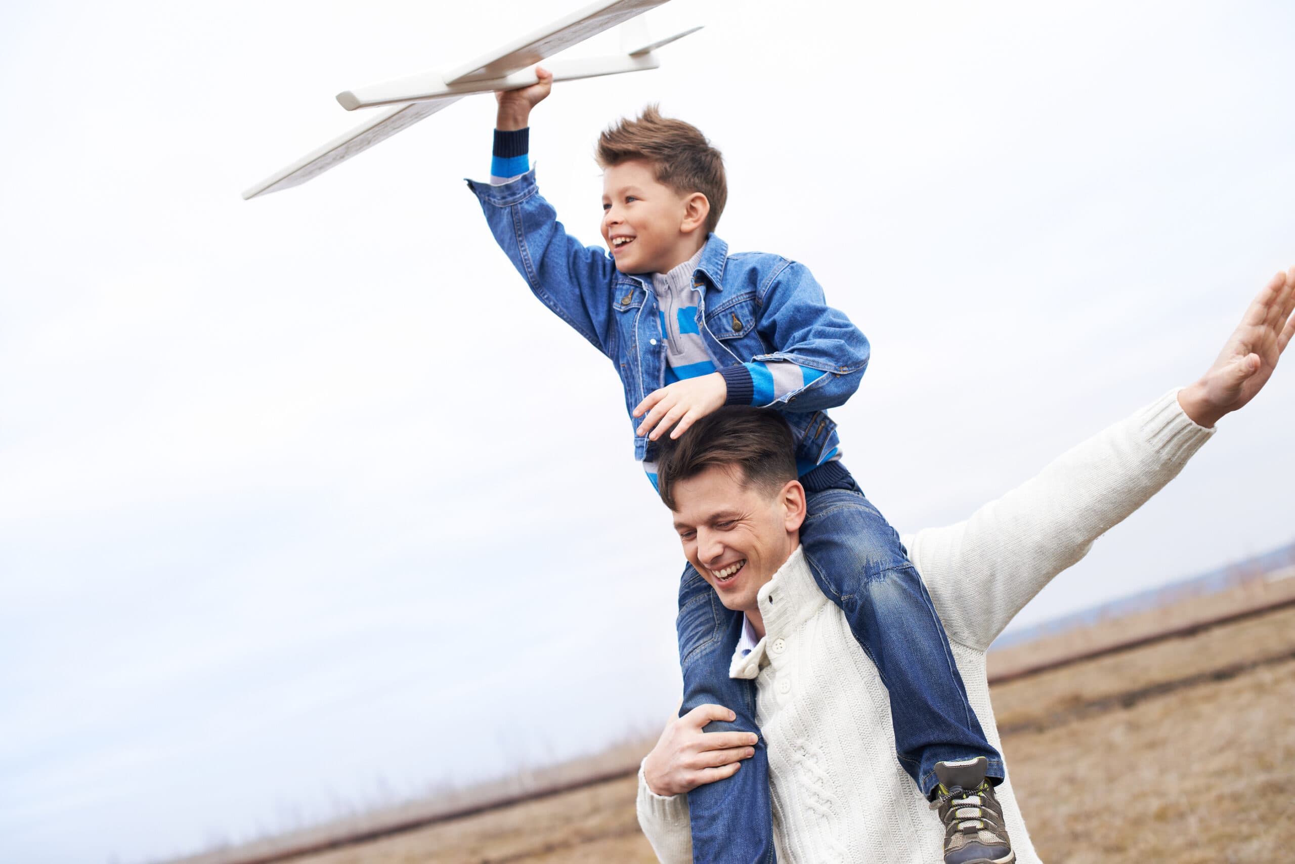 Photo of happy boy with toy airplane and his father playing outs A smiling man carries a happy boy on his shoulders outdoors. The boy holds a toy airplane above his head, both wearing casual clothes. The background is a cloudy sky and an open field.