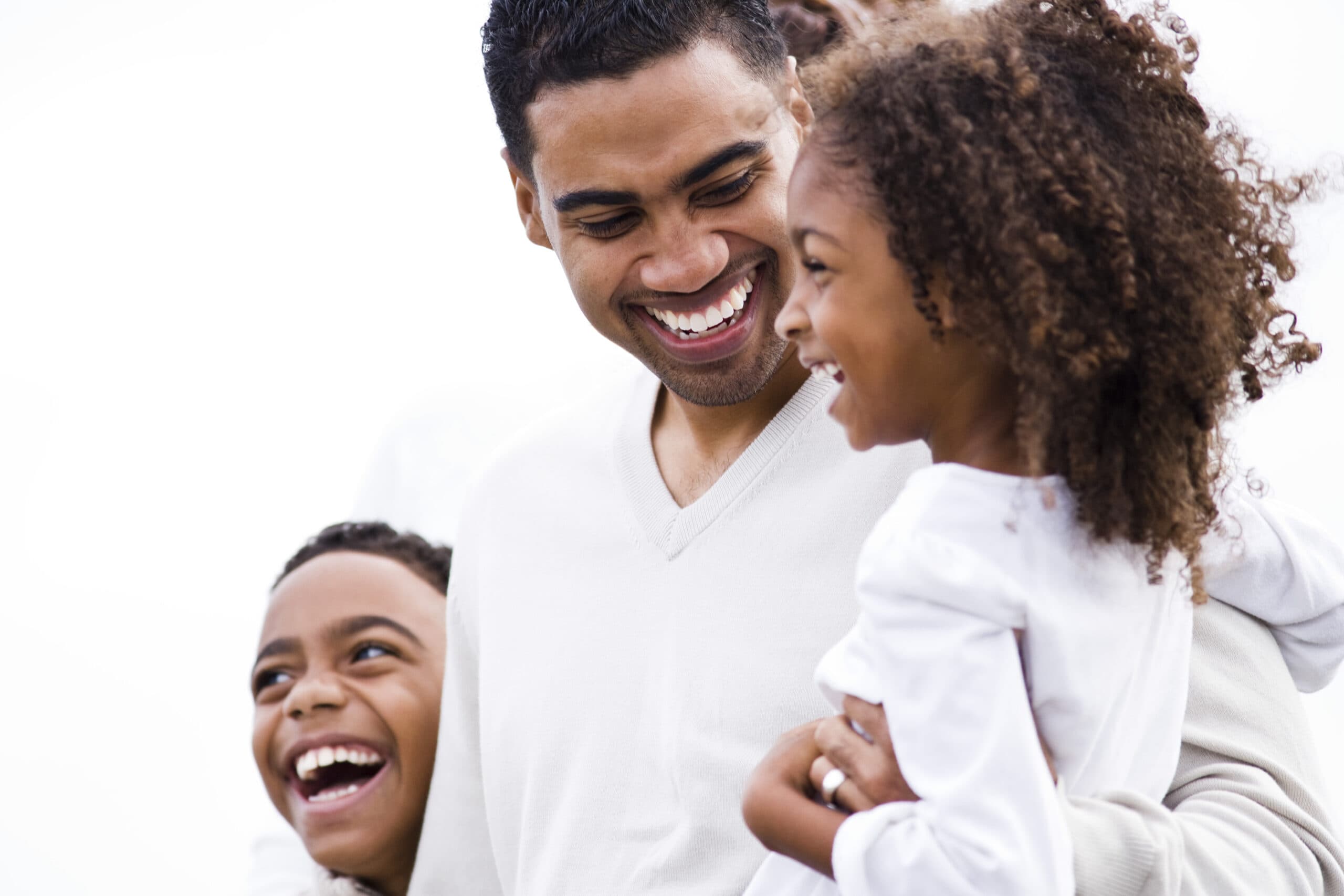 Parents Like Us A smiling man hugs two laughing children, all wearing light-colored clothes, against a bright background. They appear happy and are enjoying time together.