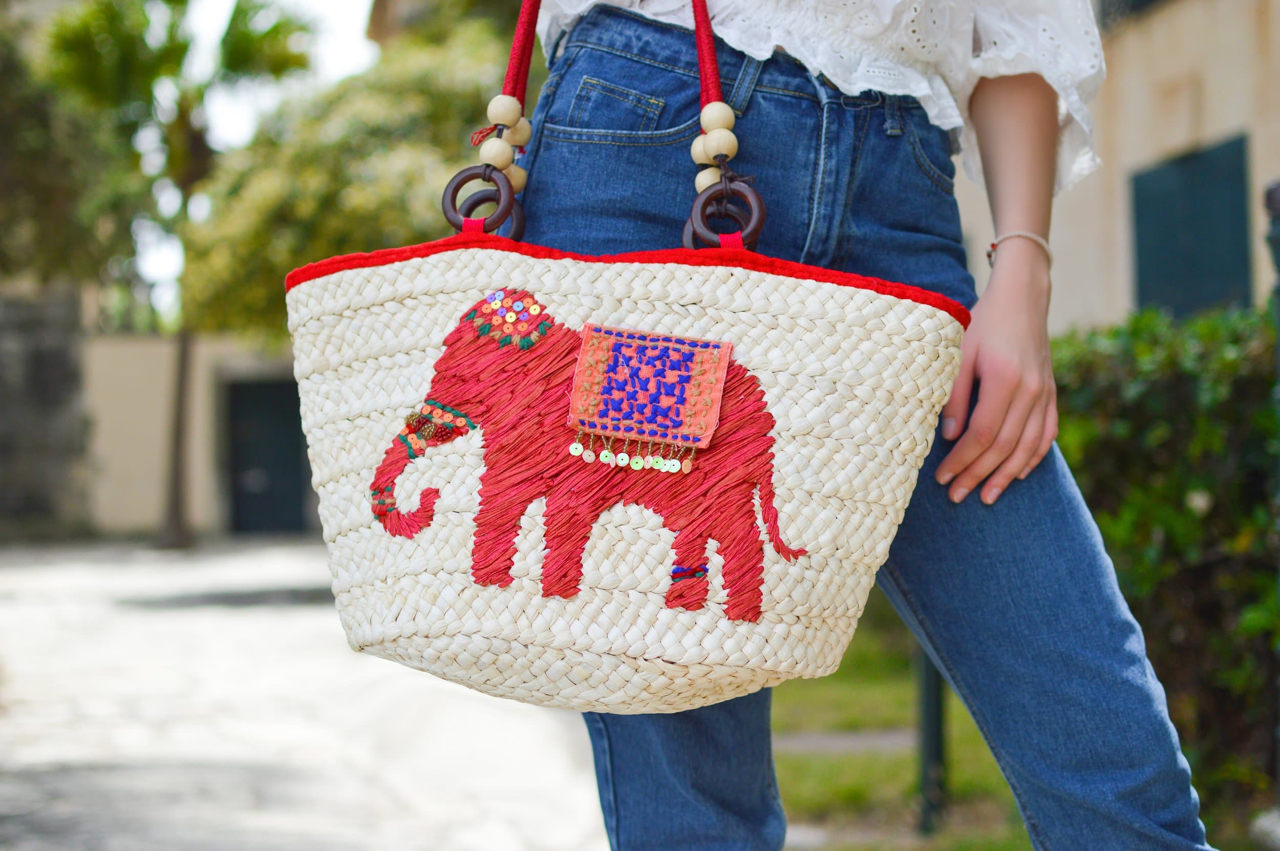 A person wearing blue jeans and a white top holds a woven straw bag decorated with a red elephant design and colorful embellishments. The background shows an outdoor setting with greenery and buildings.