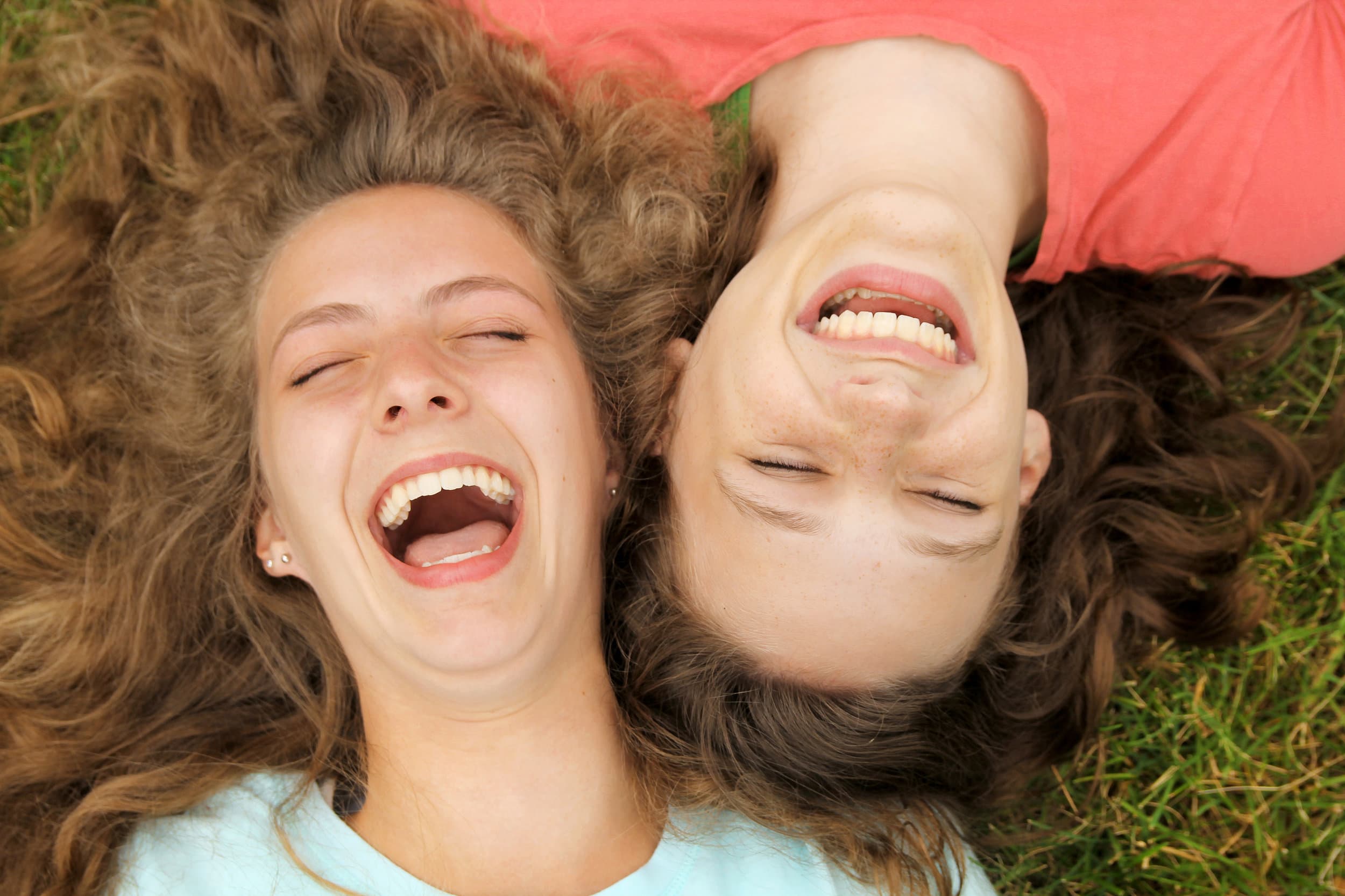 attachment-5cbe06d20d92974bf0b22440 Two young people with long, curly hair are lying on grass, heads close together, both smiling and laughing joyfully while looking up.