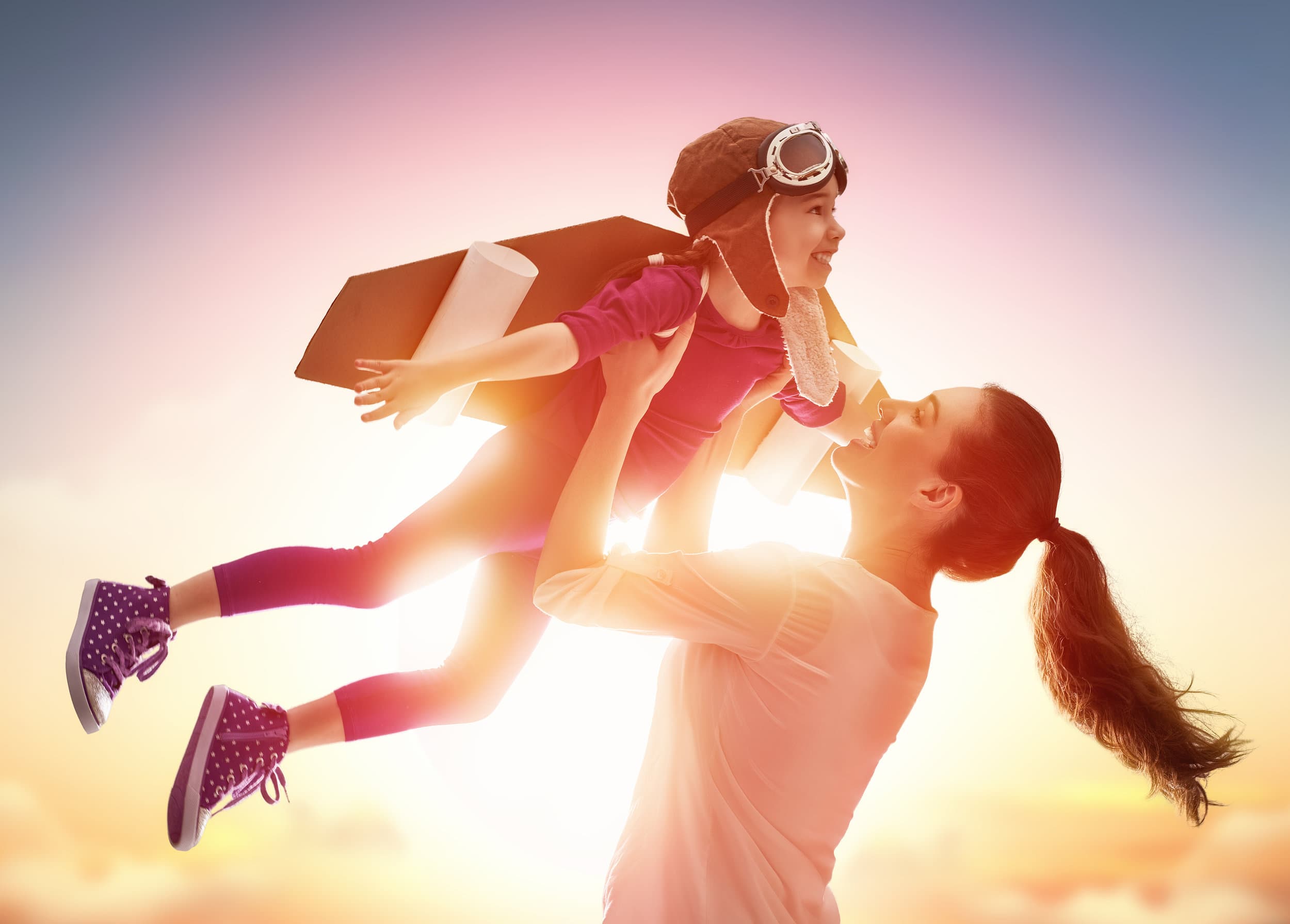 attachment-5cab57136e9a7f4f2c4ec3ef A woman lifts a smiling child into the air at sunset. The child wears a toy jetpack, aviator hat, and goggles, pretending to fly while the woman smiles up at them. Both are backlit by a warm, glowing sky.