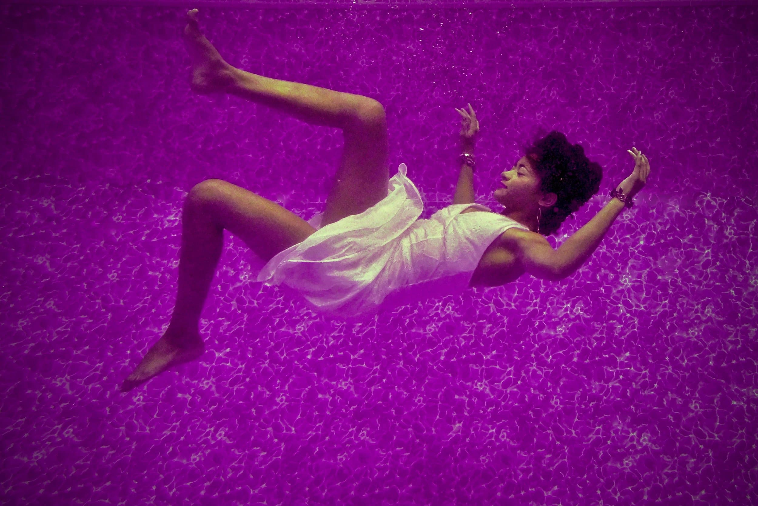 attachment-5d0a63704567f00001d7ec8f A woman in a white dress floats underwater in a swimming pool with a purple tint, arms and legs bent, looking upwards, surrounded by shimmering patterns on the pool floor.