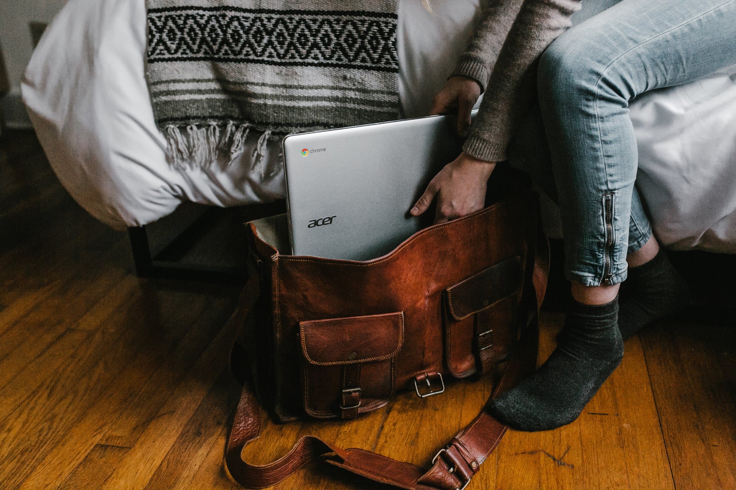 attachment-5d8c8e87c404a769007a4a87 A person sits on the edge of a bed, placing a silver Acer Chromebook into a brown leather satchel on a wooden floor. Only their hands and legs are visible.