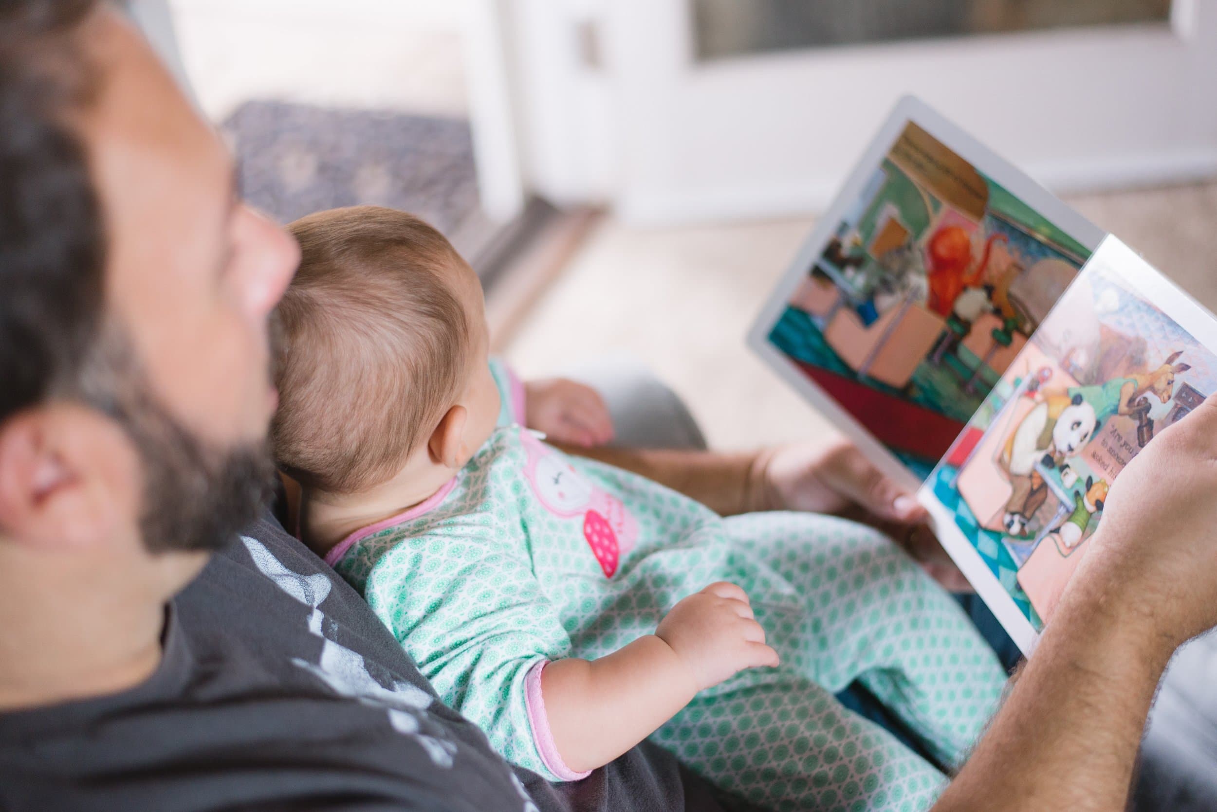 attachment-5dc461680af3a6323cb8dbfe A man and a baby sit close together as the man reads a colorful picture book to the baby, who is wearing green patterned pajamas. The scene appears cozy and intimate, with natural light in the background.