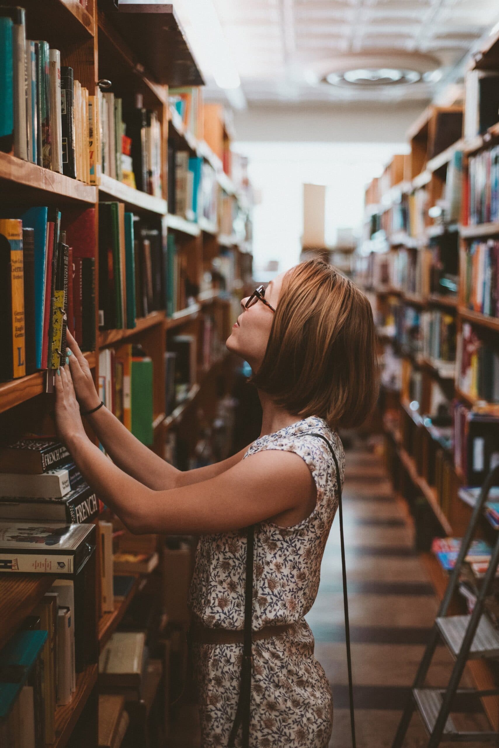 attachment-5e3851d00c38b93e1477b665 A woman wearing glasses and a sleeveless floral outfit browses books on a tall shelf in a library or bookstore, surrounded by rows of bookshelves.