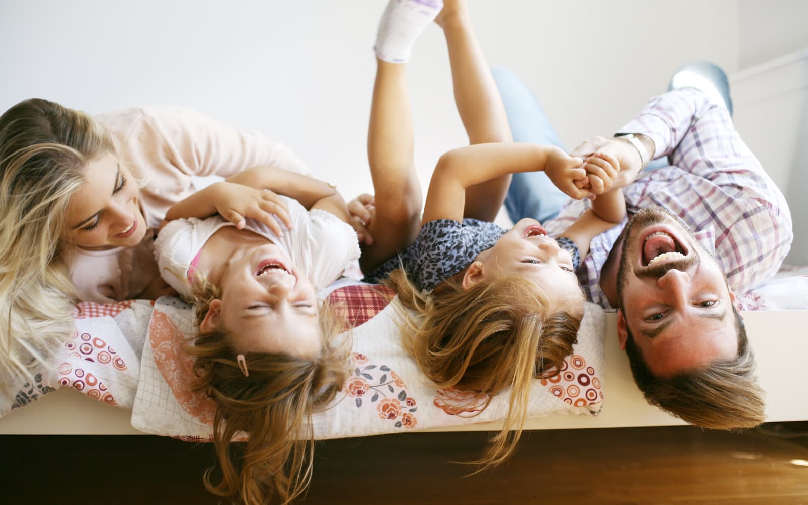 1 A family of four lies on a bed, laughing and playing together. Two young girls are in the middle, with their parents on each side. All are smiling and appear joyful, sharing a playful, happy moment.