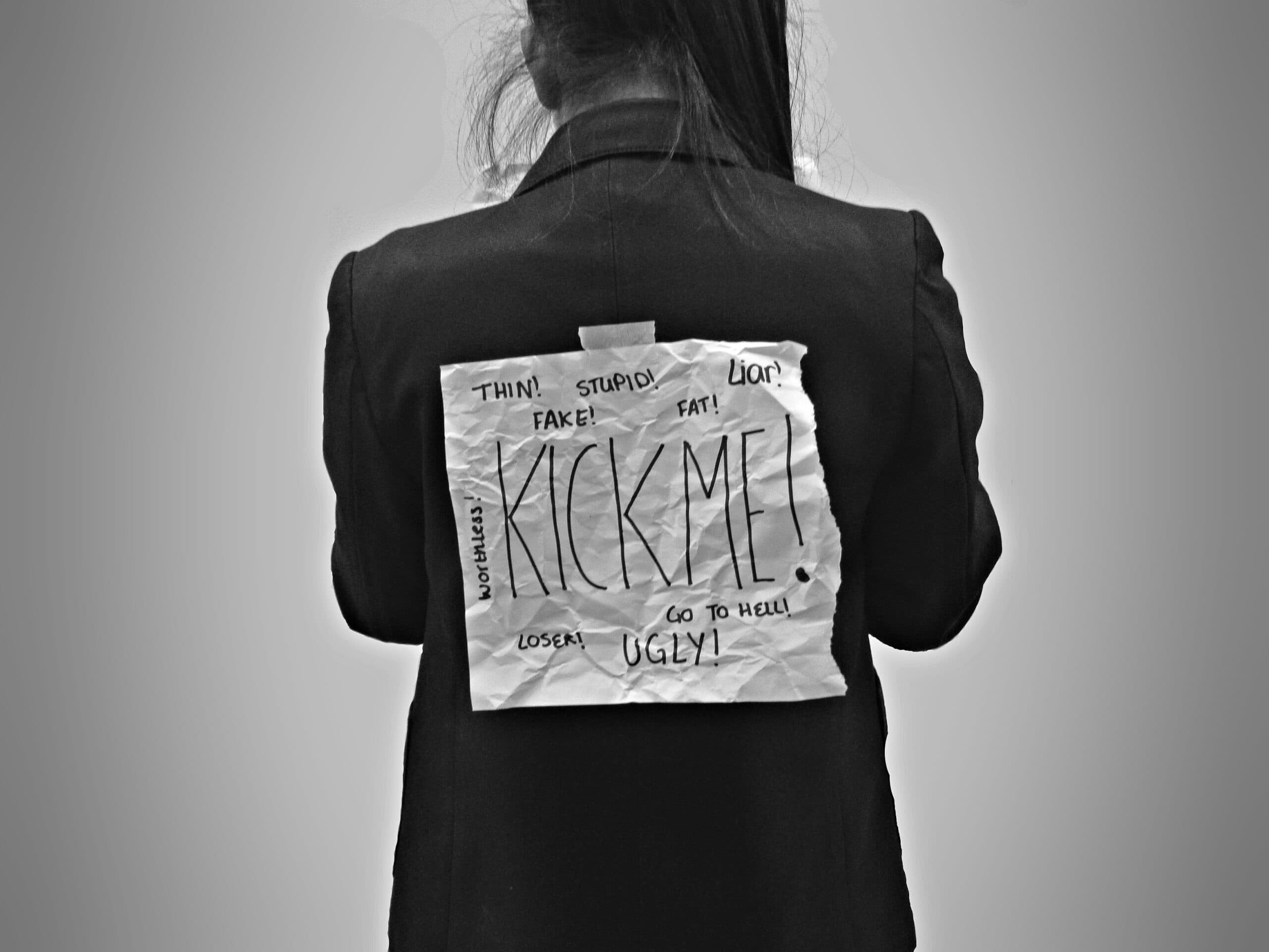 kick me ilayza-macayan A person in a dark jacket has a crumpled paper taped to their back with “KICK ME!” and several insulting words like “stupid,” “loser,” and “ugly” written on it, symbolizing bullying.