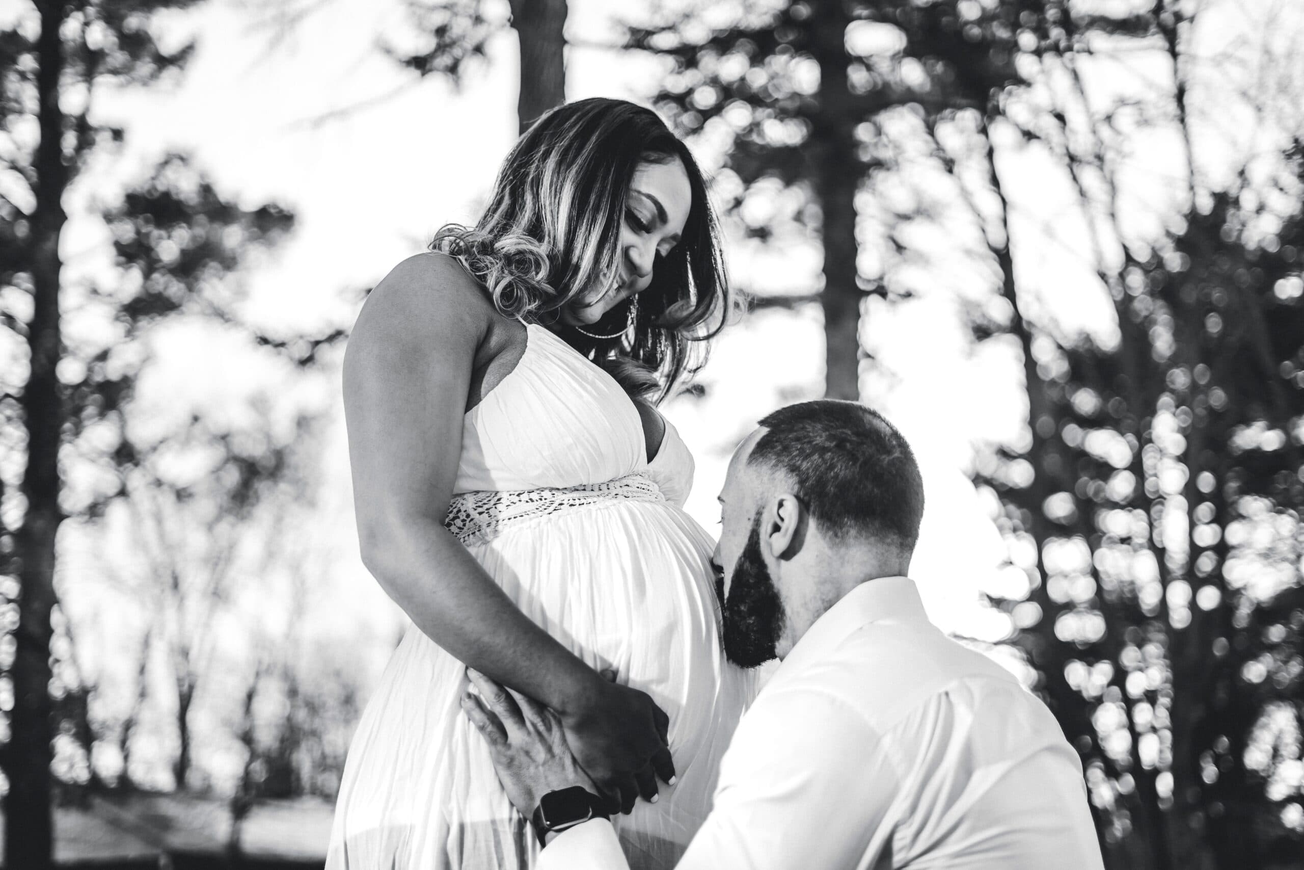 freddy-g-Z5JawooRyeI-unsplash A man kneels and kisses a pregnant womans belly as she stands, both holding hands. They are outdoors among tall trees, and the image is in black and white.