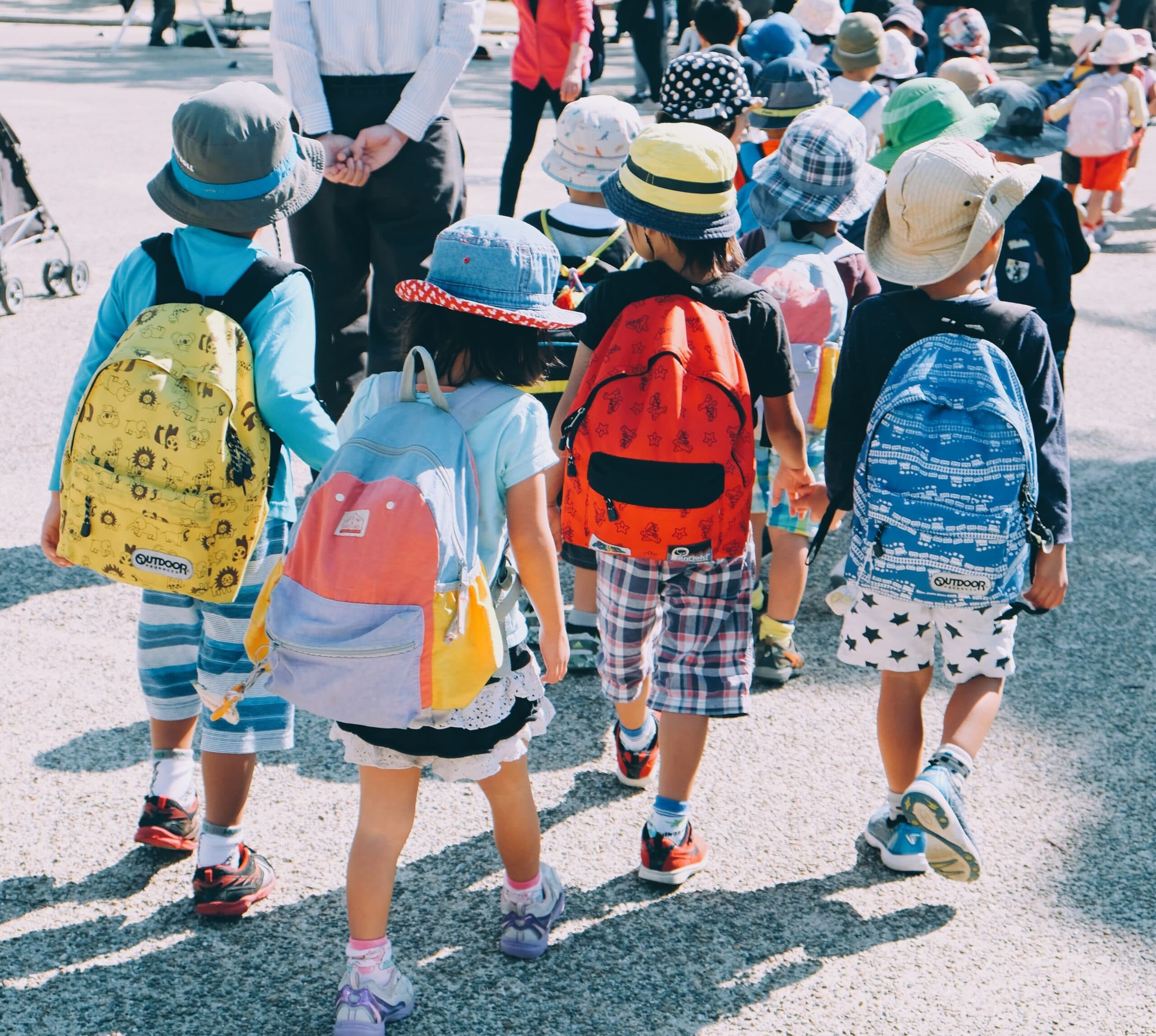note-thanun-CYlPykF-qAM-unsplash A group of young children wearing colorful hats and backpacks walk together outdoors on a sunny day, holding hands, with more children and adults visible in the background.