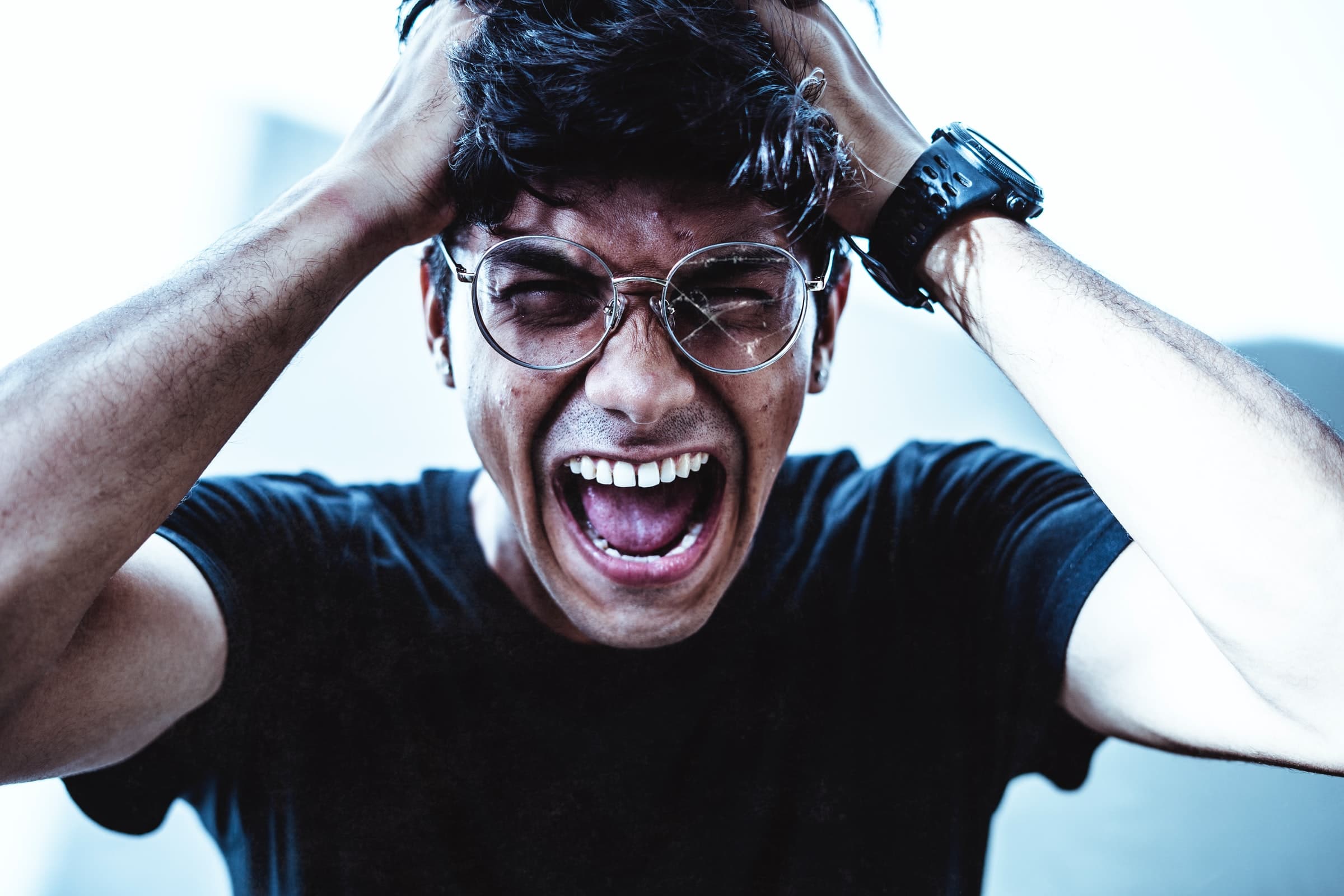 A person wearing glasses and a black t-shirt screams in frustration, pulling at their hair with both hands; their eyes are closed and mouth wide open, expressing intense emotion.