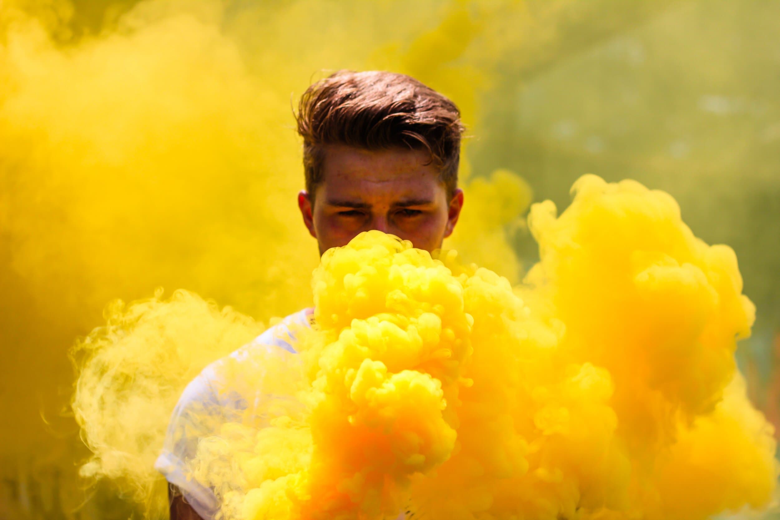 attachment-5e7fa981971cba01a86bb89c A person with light brown hair is standing outdoors, partially obscured by thick yellow smoke surrounding their face and upper body. The background is blurred with a yellow haze.