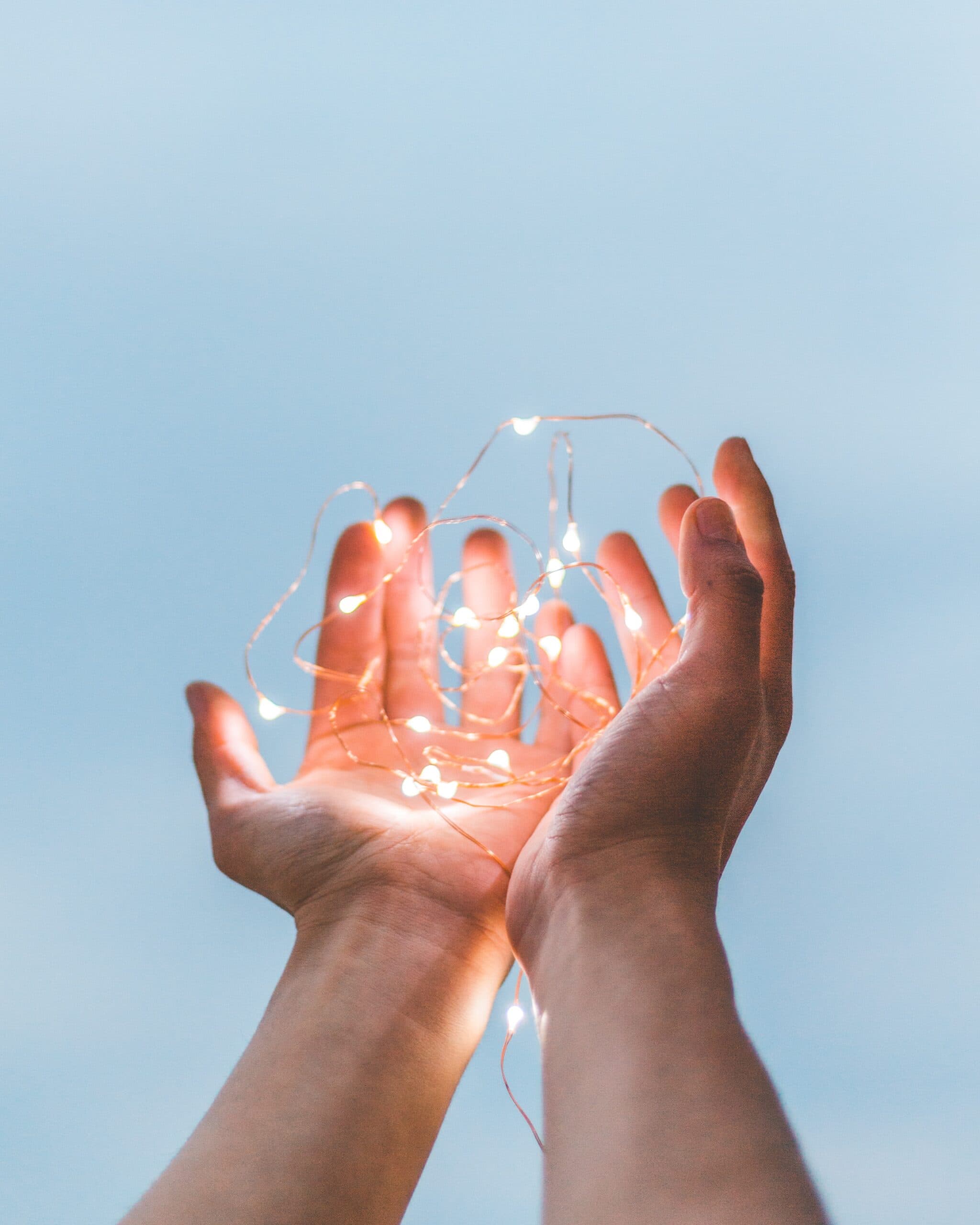 diego-ph-SZYreZsJ-fE-unsplash Two hands are held up against a pale blue sky, gently cupping a string of glowing fairy lights.