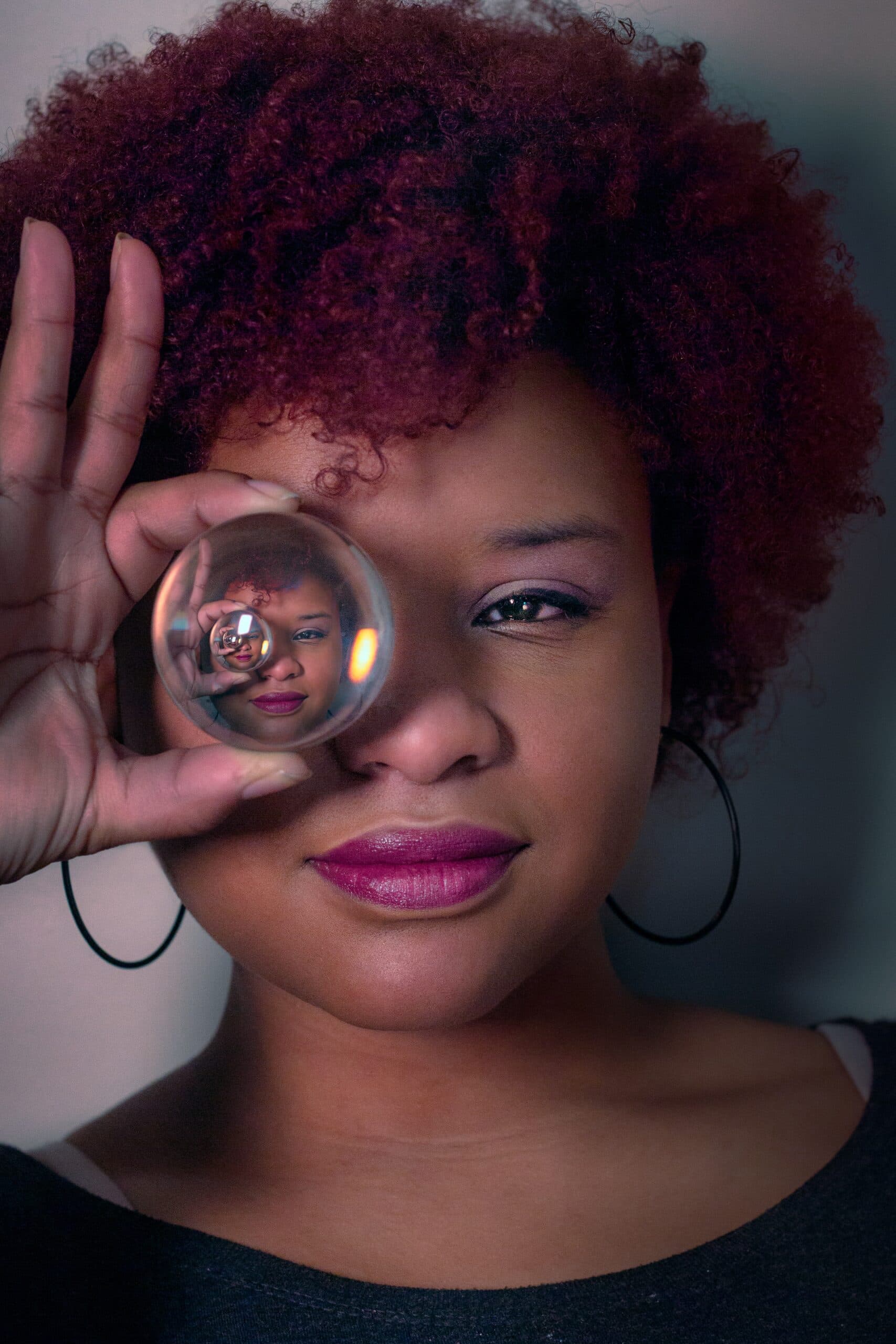 jonathan-sebastiao-qzJHFajnLCE-unsplash A person with curly red hair holds a glass sphere over one eye, reflecting their face inside the sphere. They wear hoop earrings and a dark top, and have a slight smile.