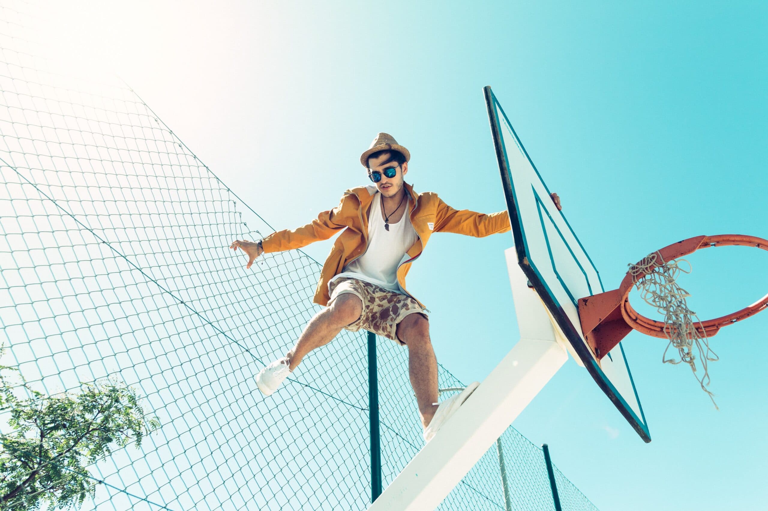 ramiro-mendes-2JMjC_jqbBk-unsplash A young man in a yellow jacket, hat, and sunglasses sits on top of a basketball hoop, holding onto the backboard, with a chain-link fence and clear blue sky in the background.