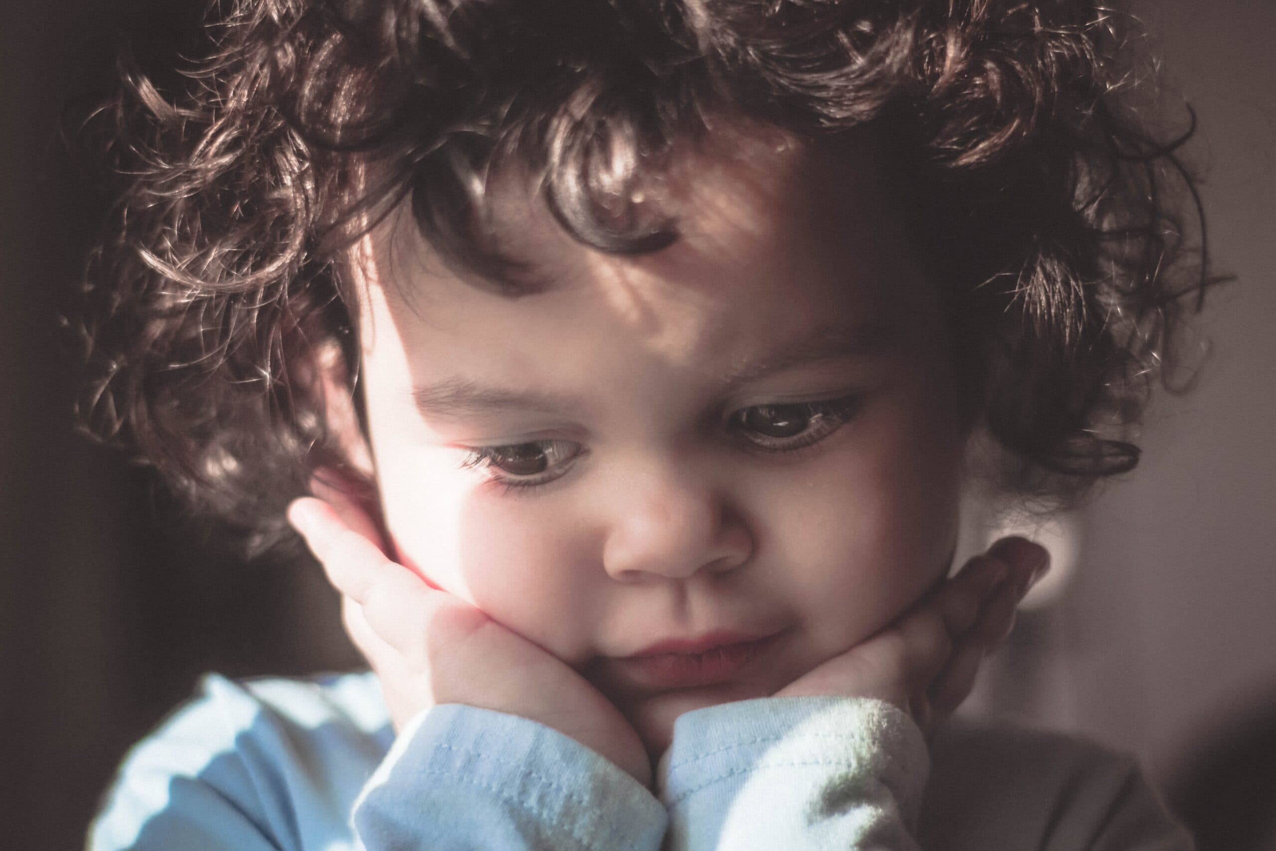 ricky-turner-anFwdlDN-zU-unsplash A young child with curly hair rests their chin on their hands, gazing downward with a thoughtful or pensive expression. Soft light highlights their face, emphasizing a calm and introspective mood.
