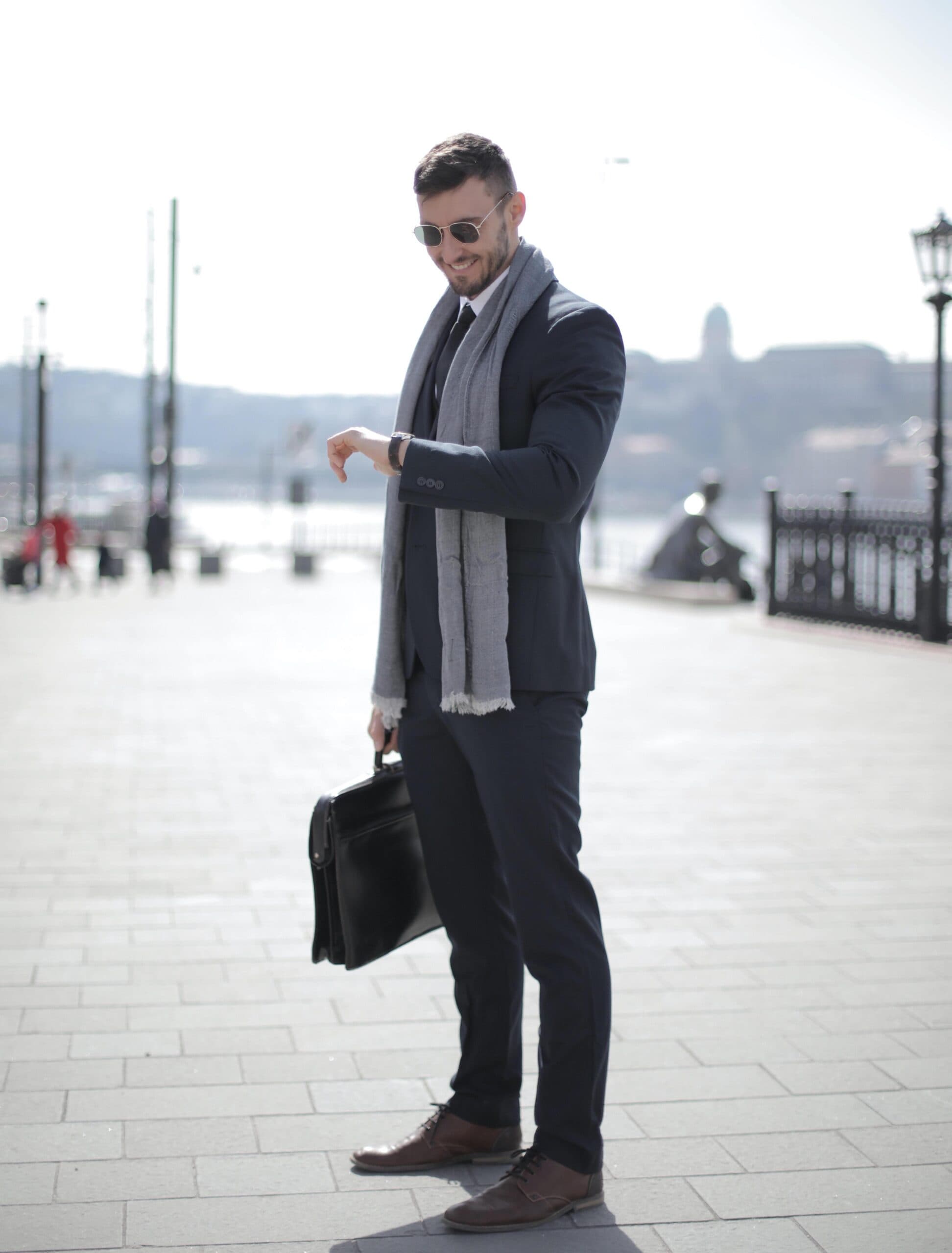 pexels-andrea-piacquadio-3778212 A man in a suit and scarf stands outdoors, holding a briefcase and looking at his watch. He is wearing sunglasses and smiling, with a cityscape and river in the background on a bright day.