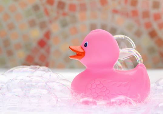 Bubble-bath-537×376 A pink rubber duck with an orange beak sits among bubbles, with a colorful, out-of-focus mosaic background.
