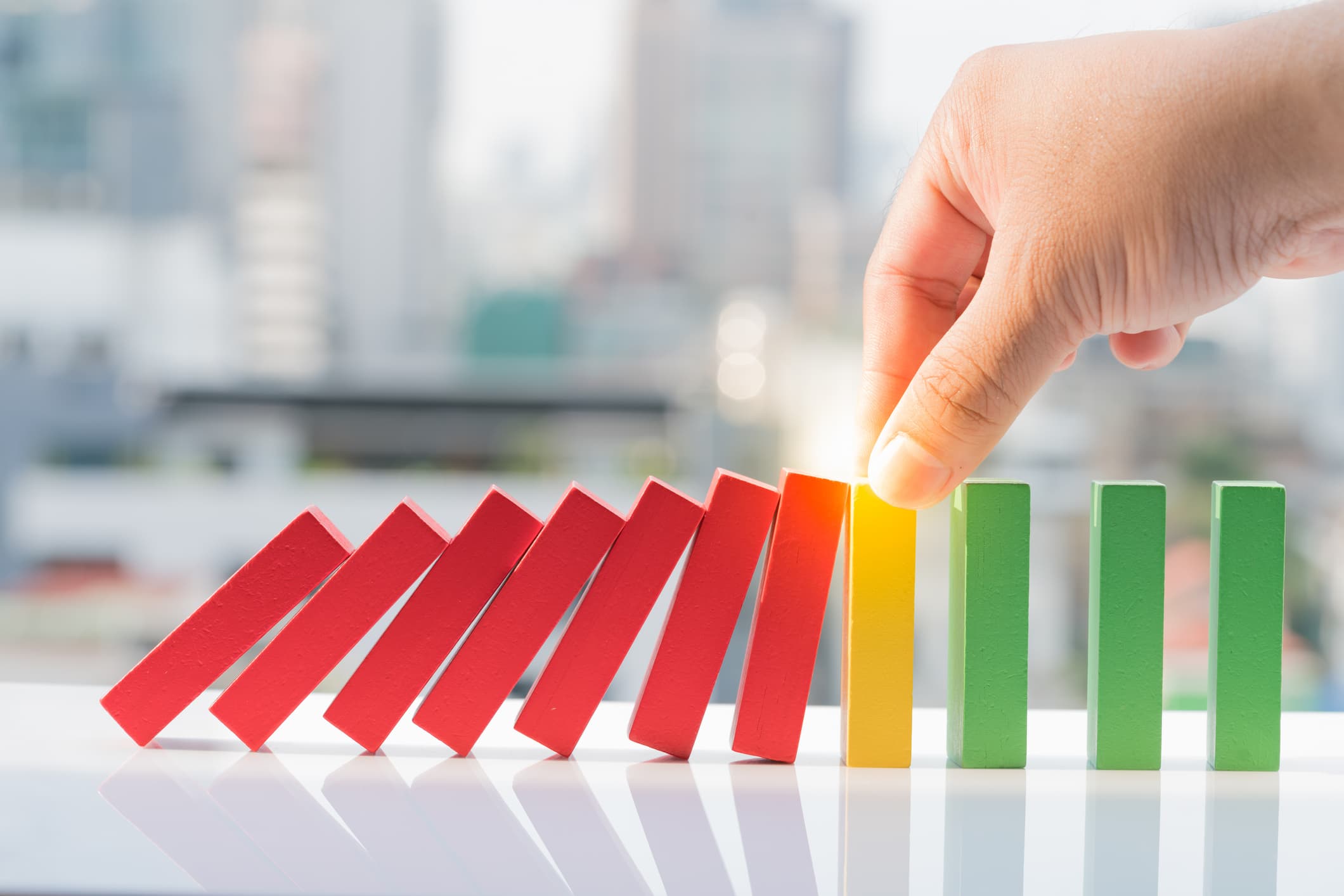 Business strategy, learning and development concepts. A hand stops a row of falling red dominoes from knocking over upright yellow and green dominoes, with a blurred cityscape in the background.