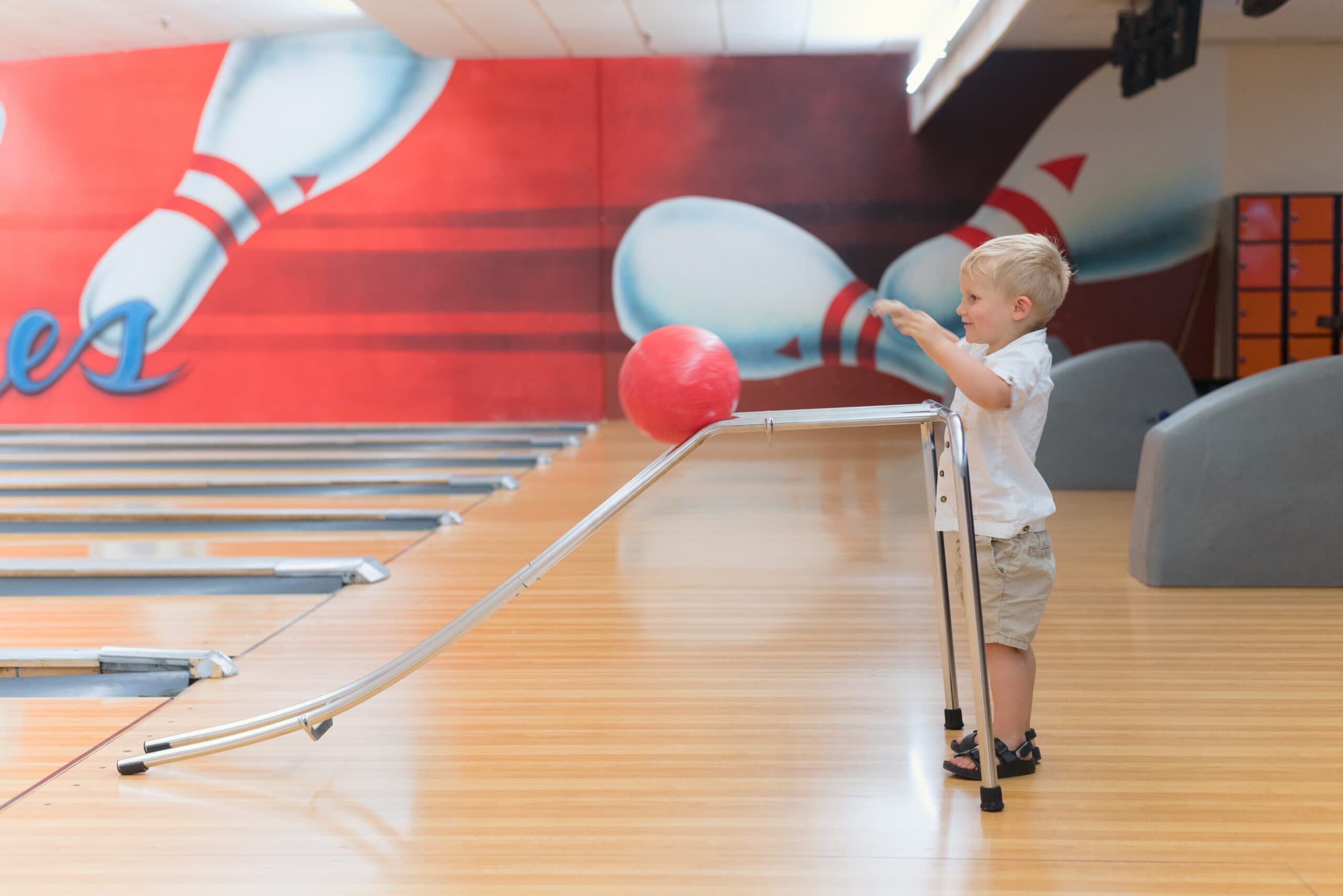 A young boy uses a ramp to roll a red bowling ball down a lane in a bowling alley with a mural of bowling pins on the wall behind him.
