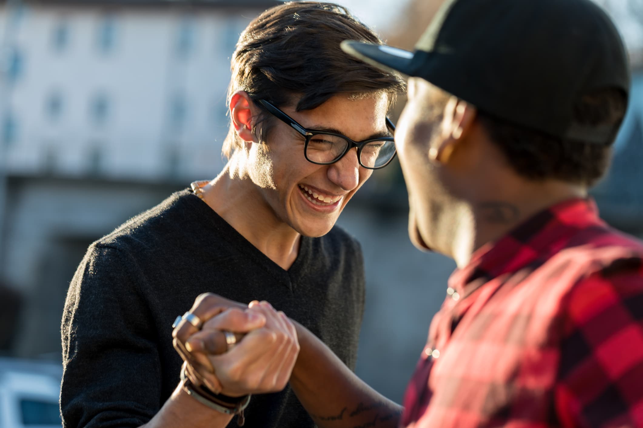 good friends joking about their secret handshake Two young men smile and laugh while shaking hands outdoors. One wears glasses and a black sweater, the other wears a red plaid shirt and a black cap. The background is softly blurred.