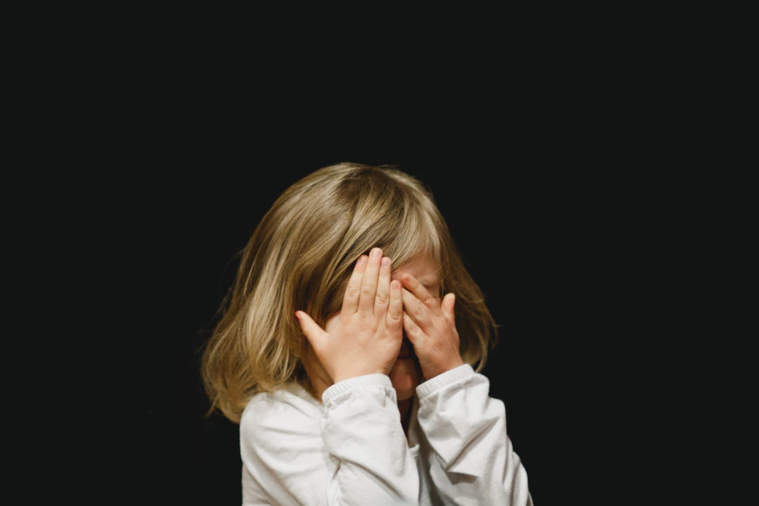 caleb-woods-VZILDYoqn_U-unsplash A child with shoulder-length blonde hair wearing a white shirt covers their face with both hands against a plain black background.