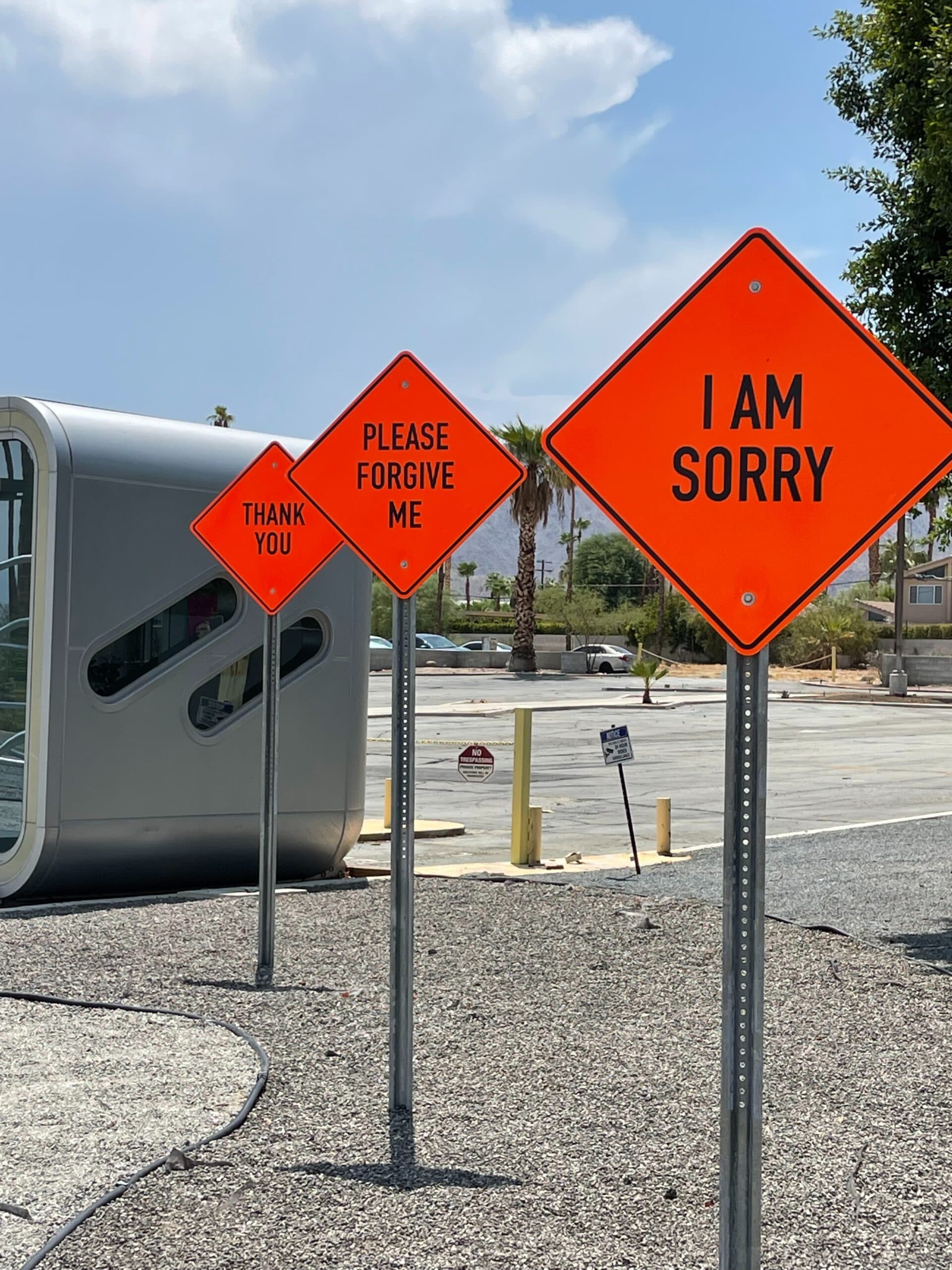 mark-tulin-L7TkIKc0XnU-unsplash Three orange diamond-shaped road signs read THANK YOU, PLEASE FORGIVE ME, and I AM SORRY. They stand in a row beside a modern building and an empty parking lot on a sunny day.