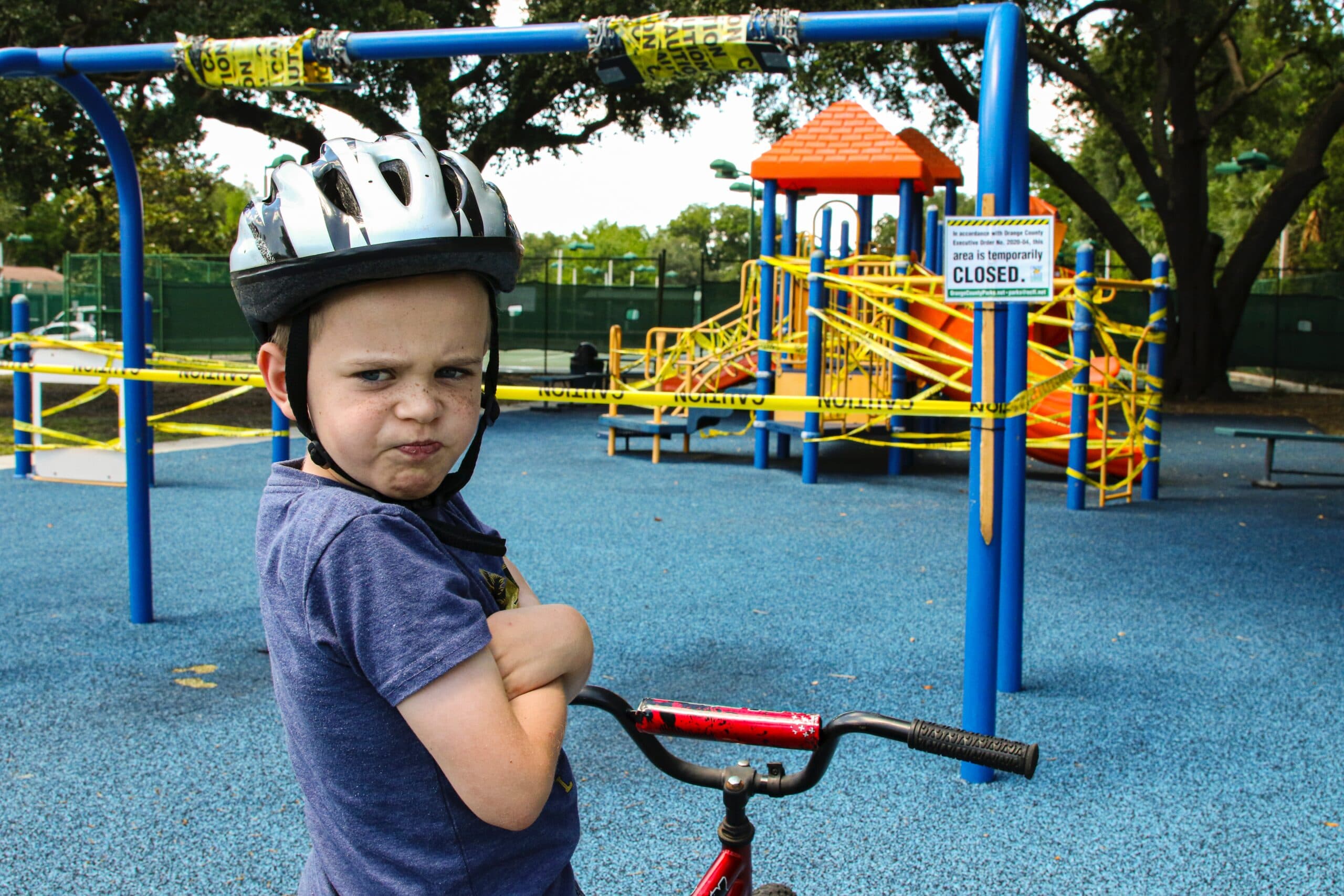mick-haupt-cDjVQiBxiMc-unsplash A young boy wearing a bike helmet pouts while holding his bike at a playground closed off with caution tape and a “closed” sign, learning firsthand about the kind vs. nice difference as he faces the restrictions. The playground equipment is visible in the background.