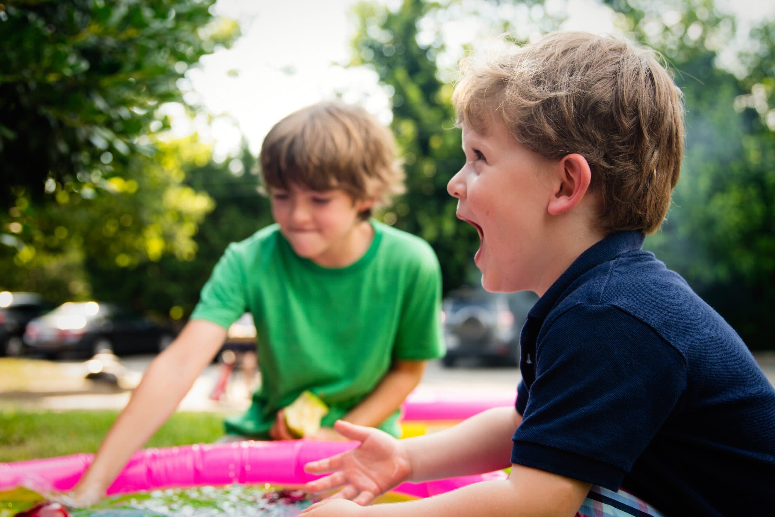 ashton-bingham-SAHBl2UpXco-unsplash Two young boys play outside next to a pink inflatable pool. Amid fatherhood challenges and ego, one boy in a green shirt reaches toward the water while the other, in a navy shirt, laughs with excitement. Trees and parked cars fill the background.