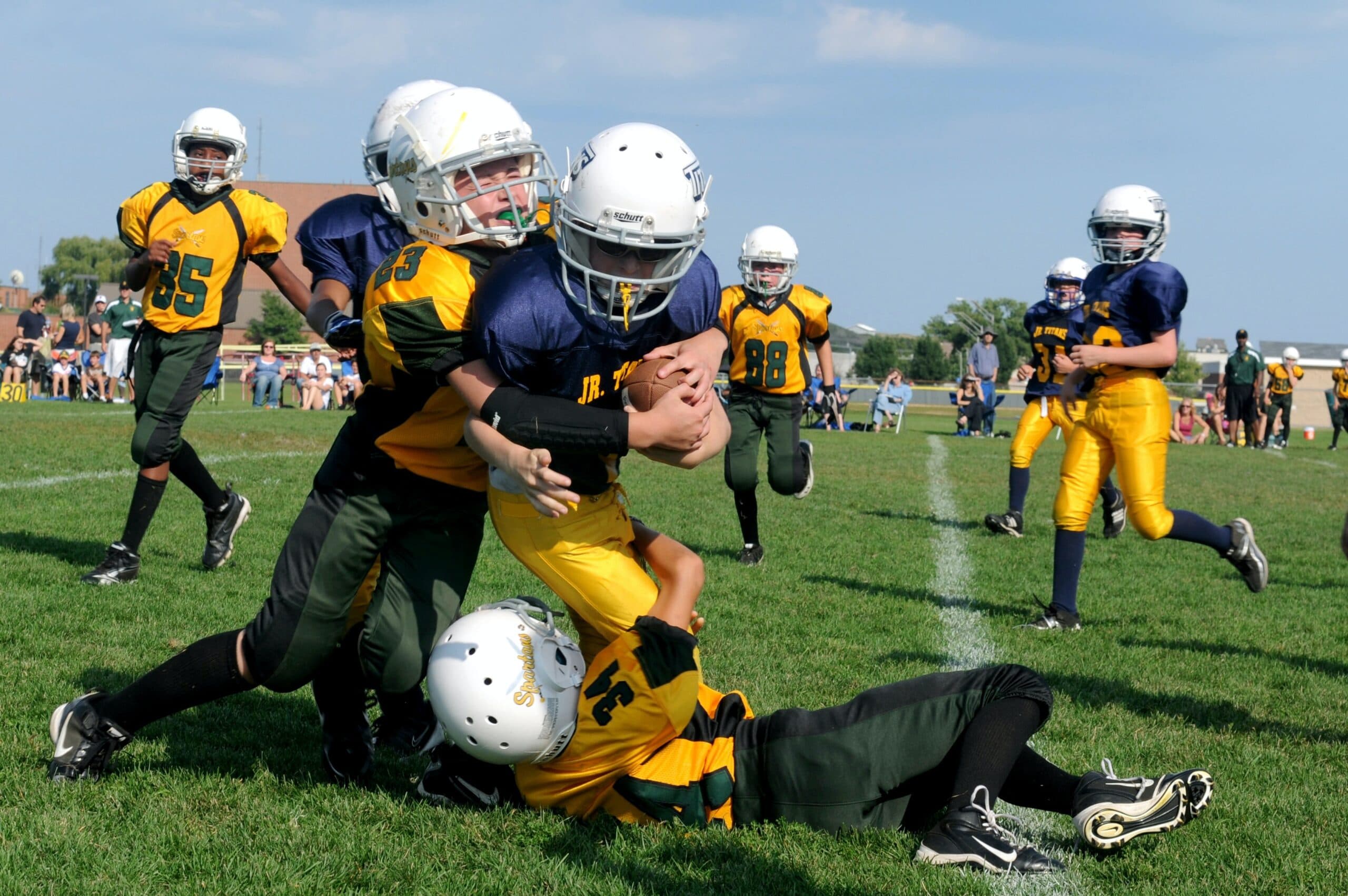 ben-hershey-nu4h8sMqees-unsplash Youth football players in helmets and uniforms tackle a player holding the ball during a game on a grassy field, breaking gender stereotypes, as other players and spectators watch in the background.