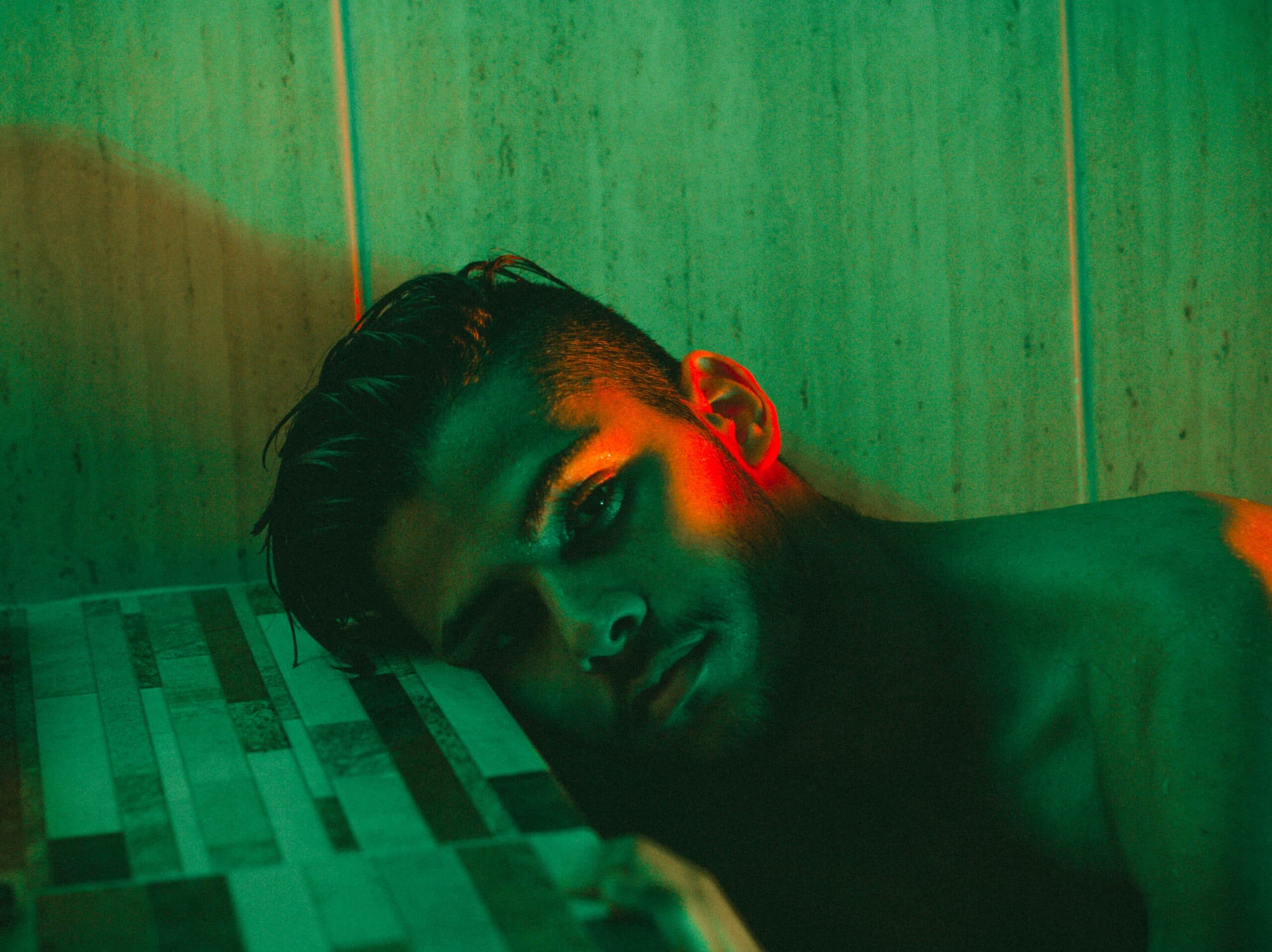 fernando-cferdophotography-5CnrCrmwwIU-unsplash A shirtless person with wet hair lies with their head on a tiled surface, illuminated by dramatic green and red lighting against a tiled wall—capturing the vulnerability of finding human connection in an urban solitude.