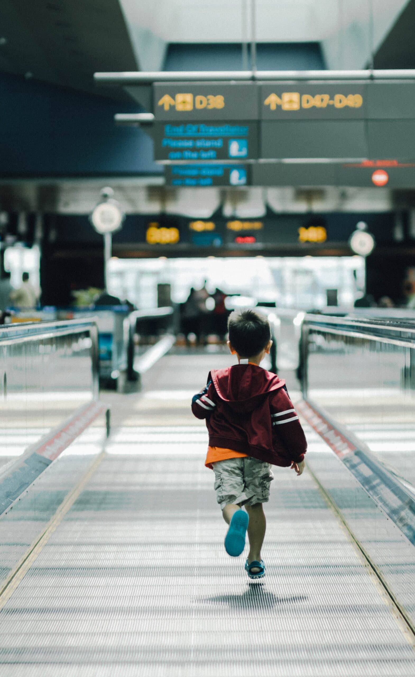 hanson-lu-qNHbXkO3Xbc-unsplash A young child in a red jacket walks alone on a moving walkway inside an airport, embodying the journey of embracing failure and growth, with signs for gates and blurry travelers visible in the background.