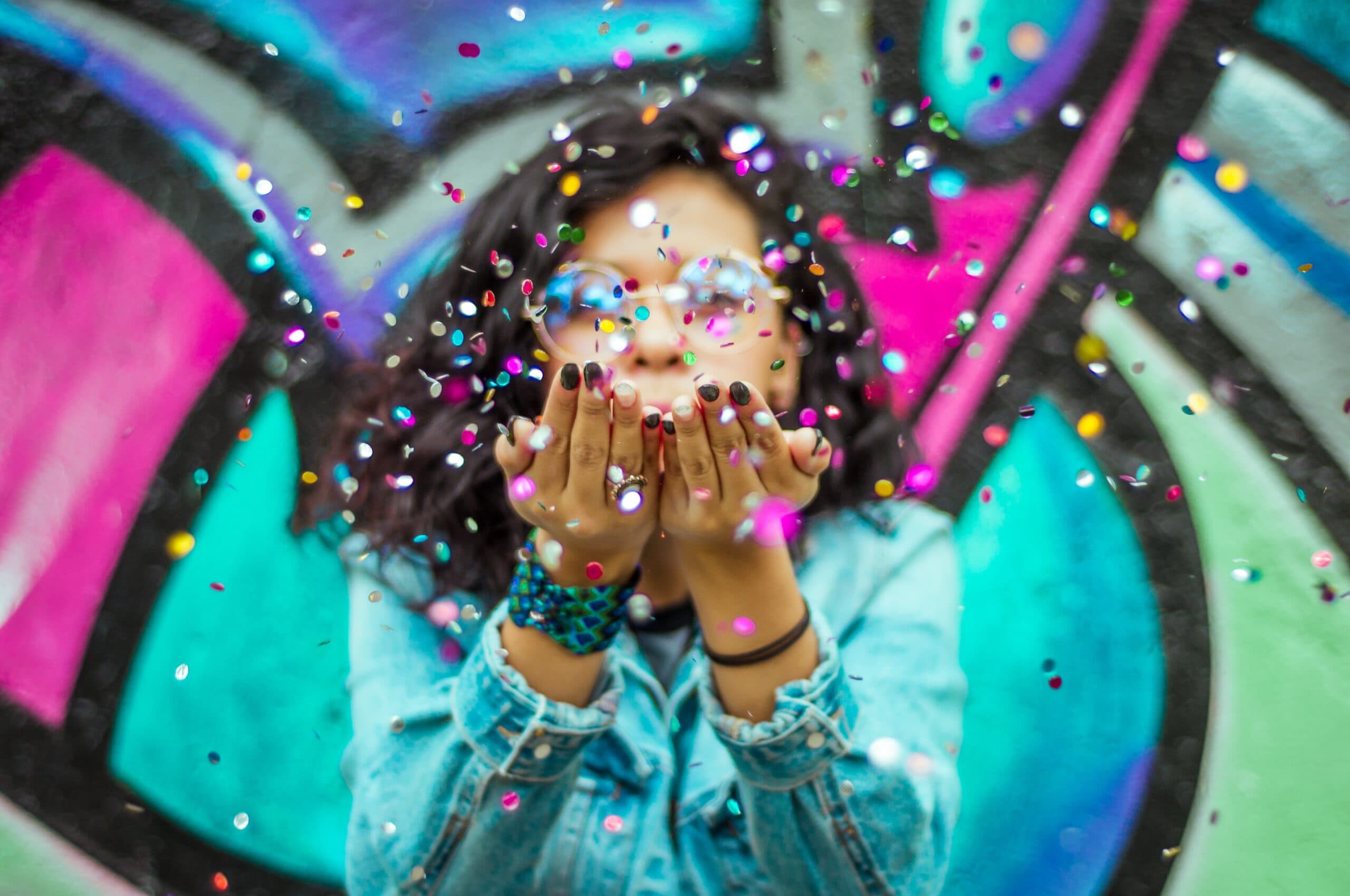 hugo-ruiz-e2pVrE1PYzs-unsplash A person wearing glasses and a denim jacket blows colorful confetti toward the camera, with a vibrant graffiti wall in the background—perfect inspiration to make your own free Father's Day card.
