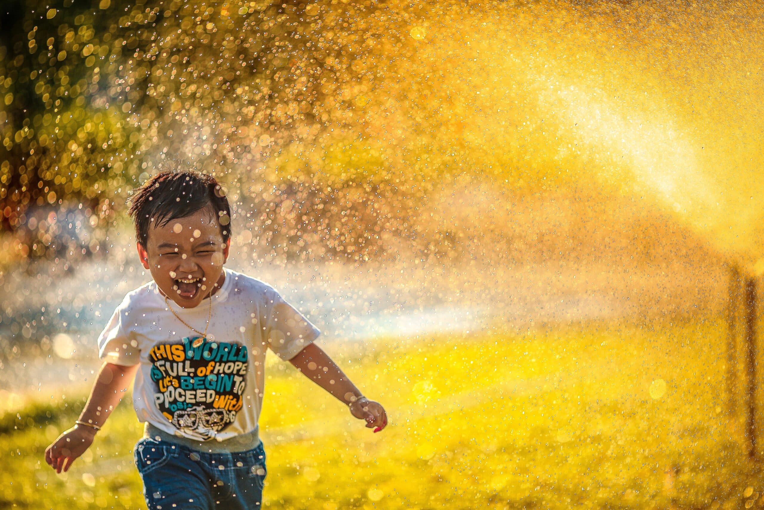 mi-pham-FtZL0r4DZYk-unsplash A young child laughs joyfully while running through a spray of water outdoors, sunlight shining through the droplets like meditation hacks for the soul. The child wears a white t-shirt and blue shorts, every moment free from wasted efforts.