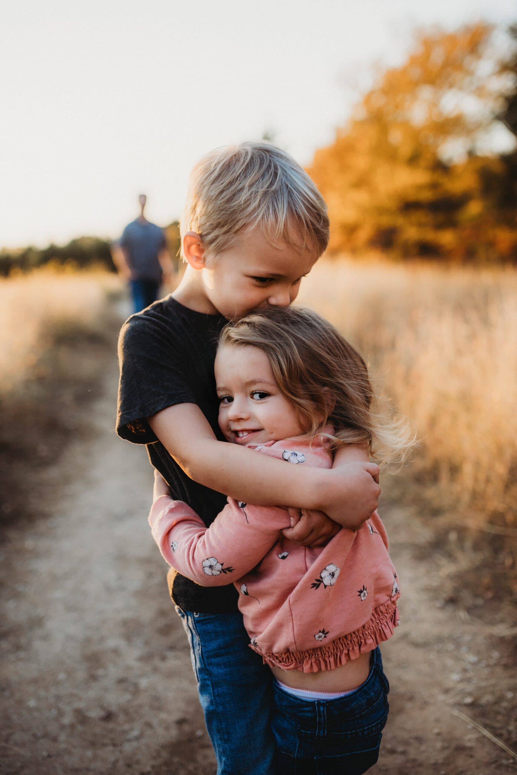 patty-brito-eHOZjZEx7u8-unsplash A young boy and a little girl hug each other on a dirt path through tall grass, embodying the power of apology. The girl smiles at the camera while the boy kisses her head, as an adult walks in the blurred background under warm evening light.