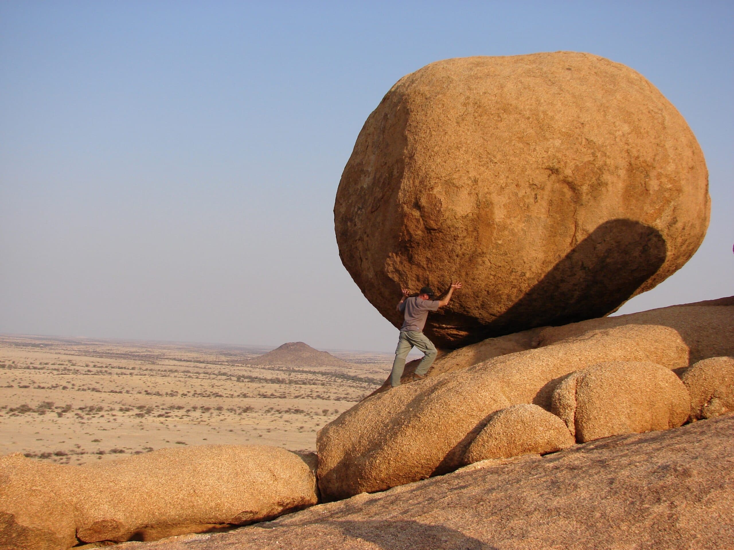 vicky-sim-mj51pZwan9c-unsplash A person appears to push a massive round boulder atop rocky terrain in a wide, arid landscape under a clear sky, symbolizing the challenge of managing change amidst difficult conditions.