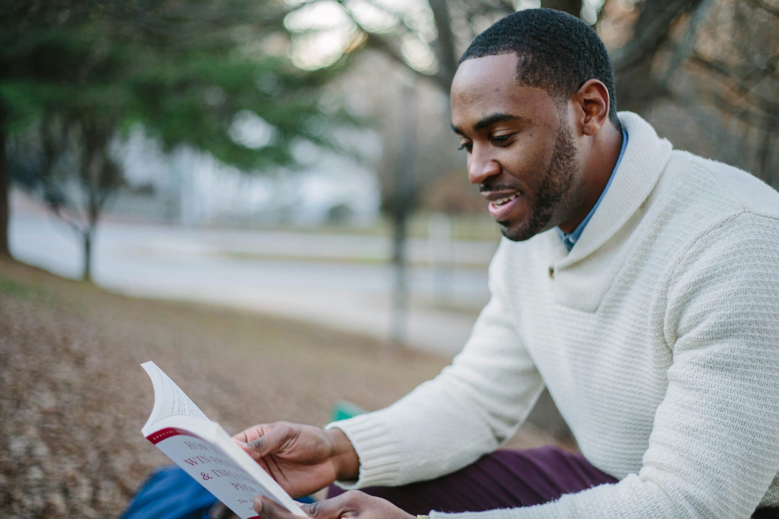 A man wearing a white sweater sits outdoors on a bench, smiling as he reads 