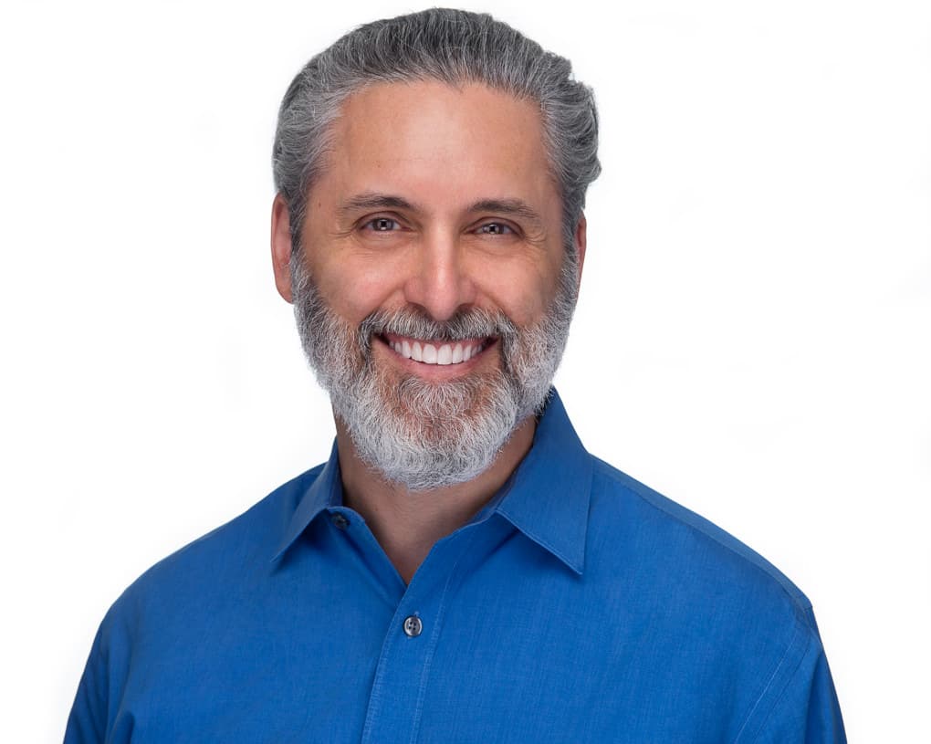 A middle-aged man with gray hair and a beard smiles warmly, wearing a blue collared shirt against a plain white background.