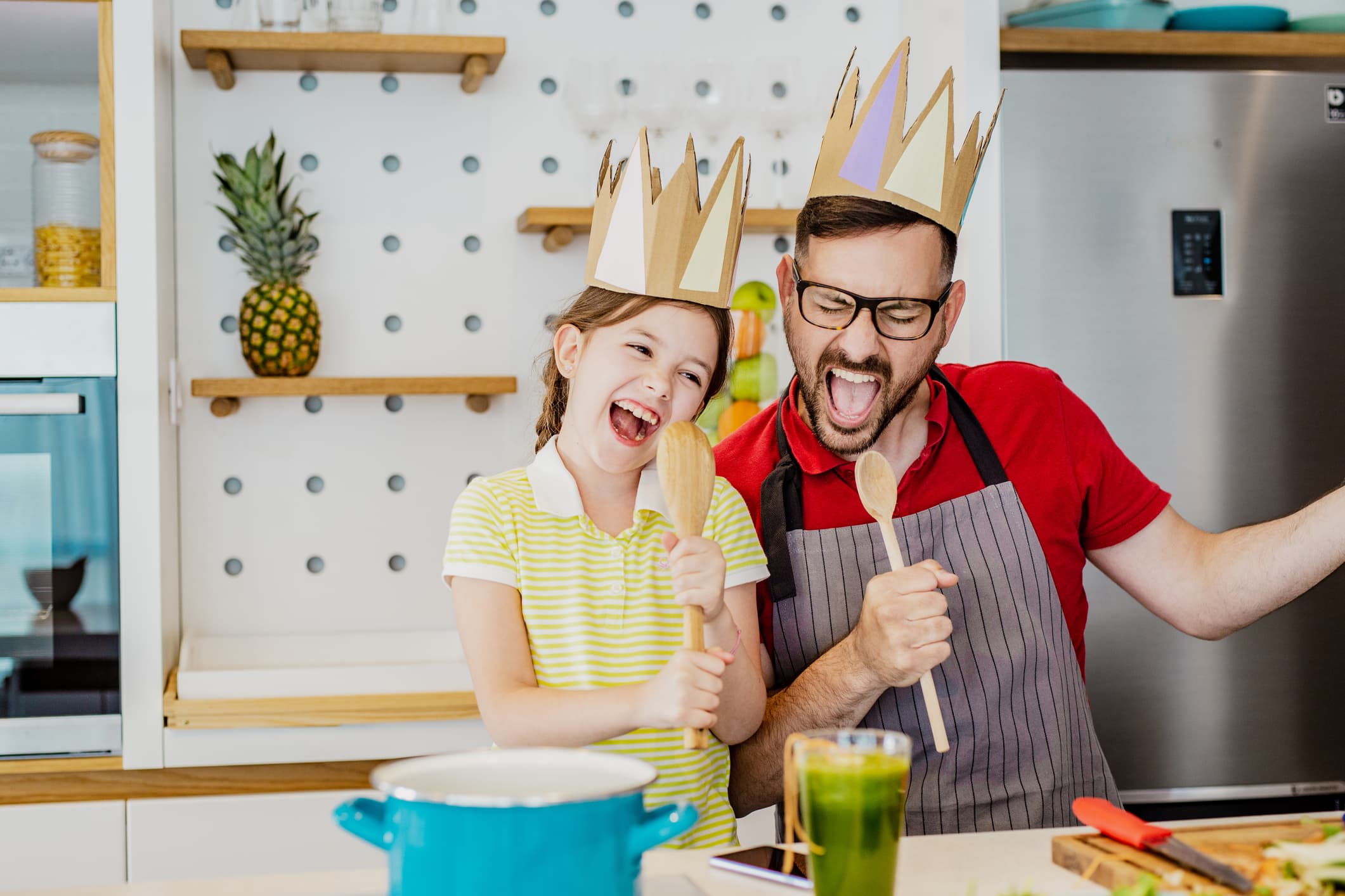Father and his little cute daughter are having fun in the kitchen while cooking A man and a girl wearing paper crowns sing and laugh together in a kitchen, holding wooden spoons as microphones. They stand next to a stove with a blue pot and a green drink on the counter.