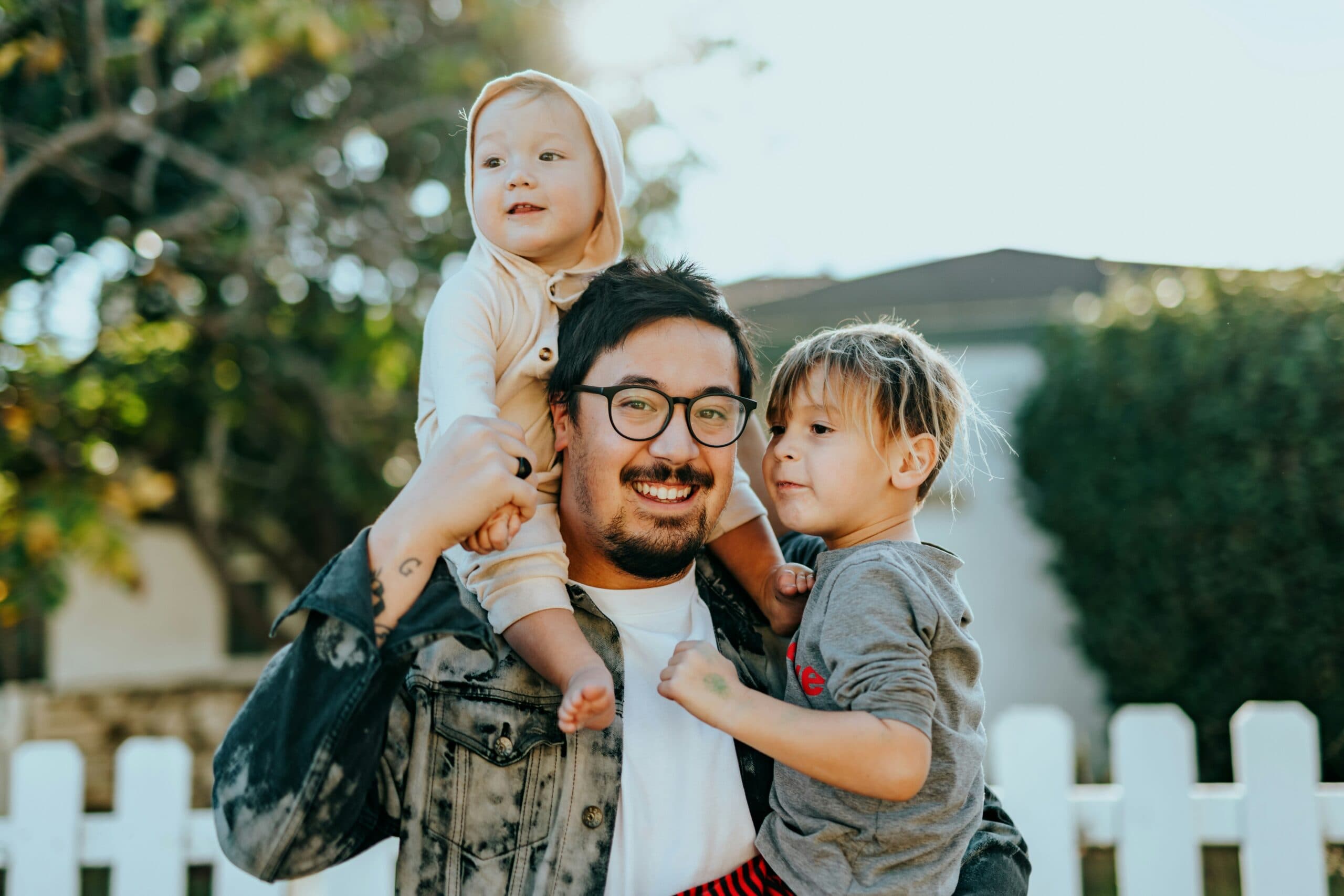 nathan-dumlao-Wr3comVZJxU-unsplash A smiling man with glasses holds a baby and a young child outdoors in front of a white picket fence, with trees and a house in the background. The baby wears a hooded outfit and the child wears a gray shirt.