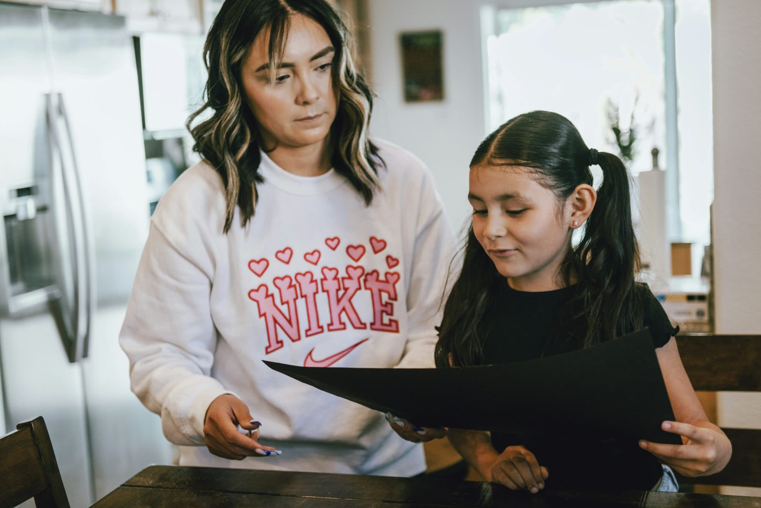 A woman in a white Nike sweatshirt stands beside a young girl with pigtails, who is holding a large black sheet of paper at a table in a bright kitchen.