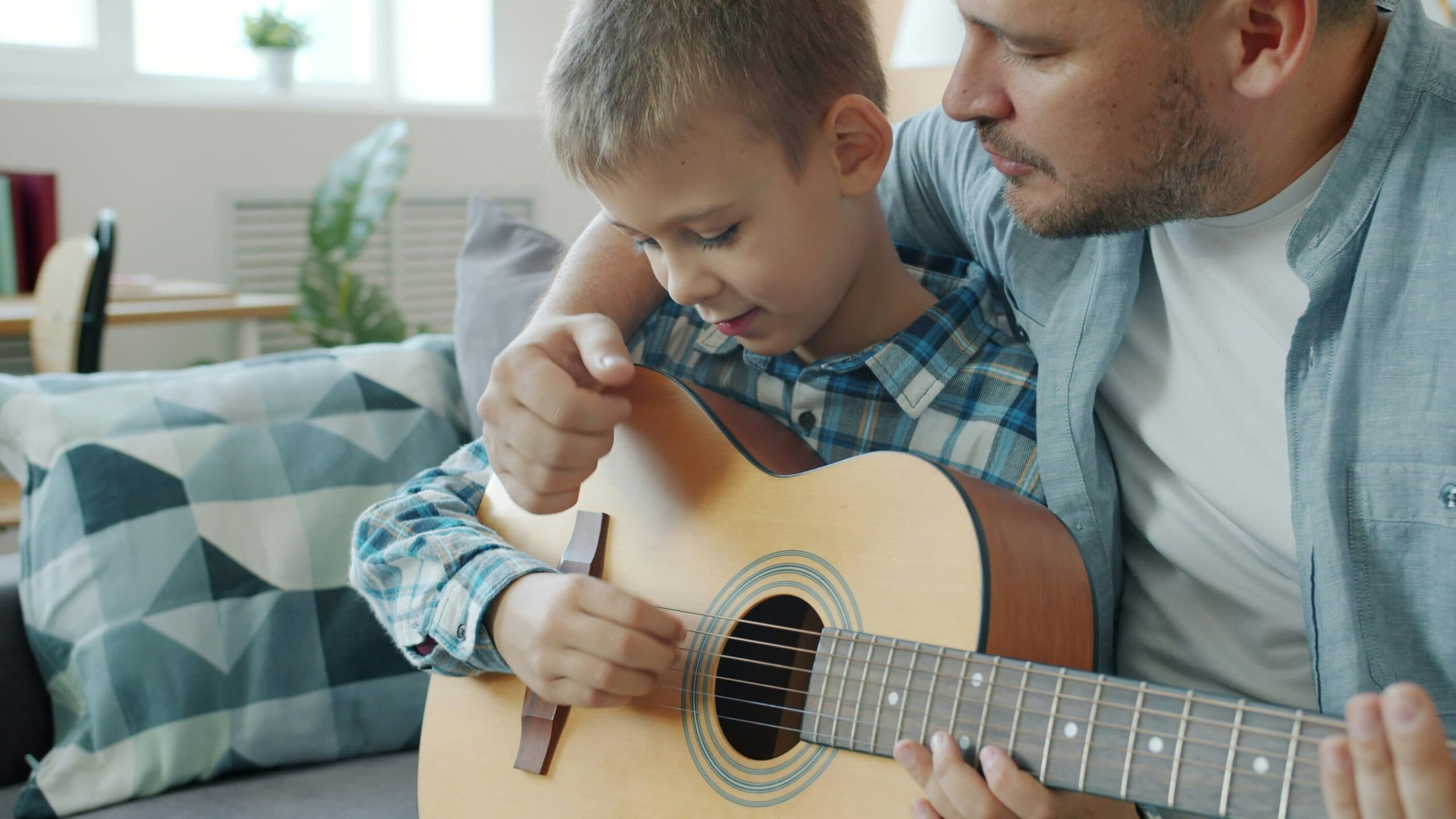 vitaly-gariev-NVWc_fyctlk-unsplash A man helps a young boy play an acoustic guitar, guiding his hand on the strings. They are sitting closely together on a couch in a bright living room with patterned pillows.
