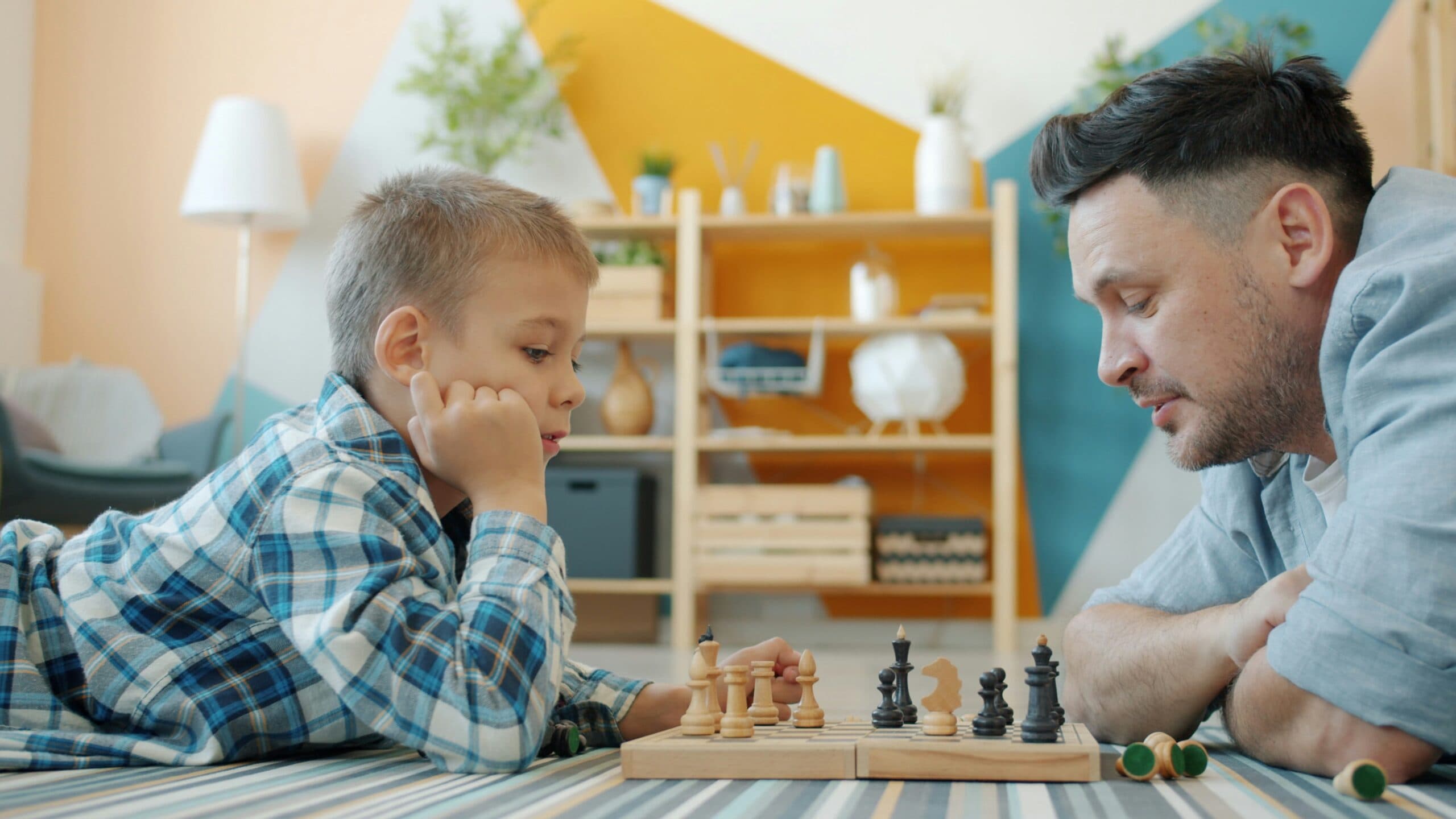 A young boy and a man lie on the floor facing each other, focused on a chess game—an engaging moment that highlights focus and strategy, much like ADHD Evaluations and Coaching support personal growth in a cozy, homey setting.