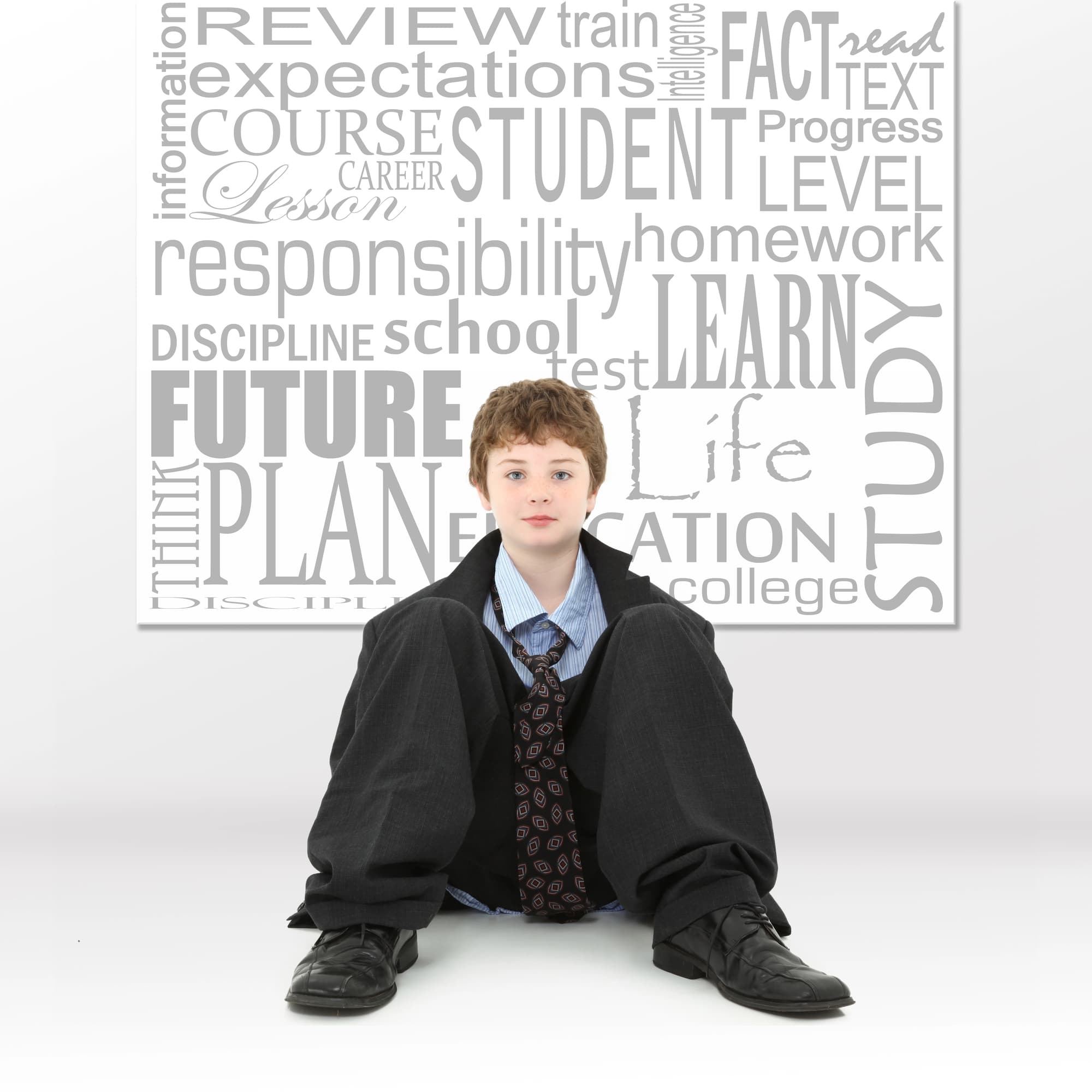A young boy in an oversized suit sits on the floor in front of a wall covered with large education-related words like future, plan, student, learn, and study.