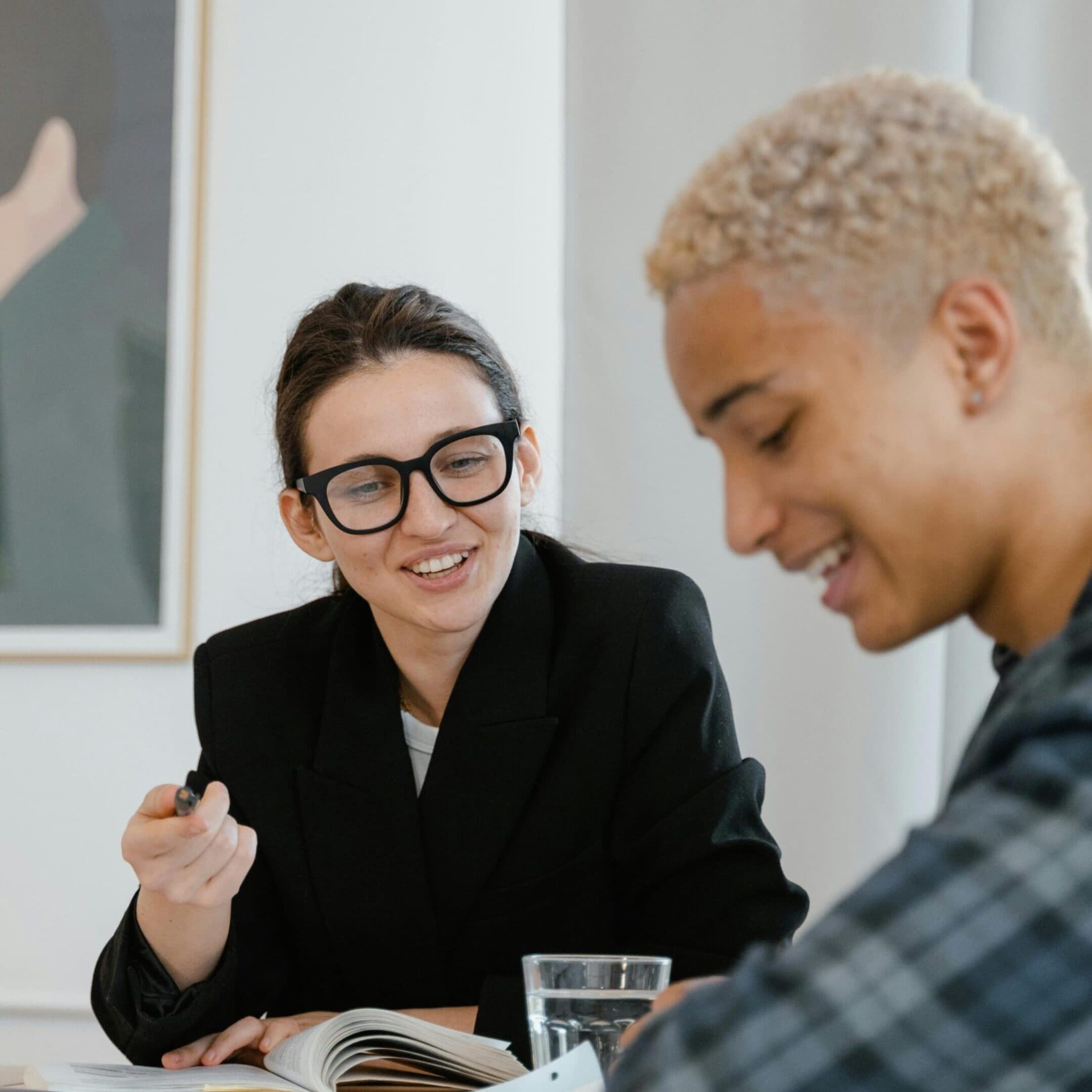 Exec Function Coaching Two people sit at a table, smiling and talking. One person wears glasses and holds a pen, while the other looks down at papers. A framed artwork is visible on the wall behind them.