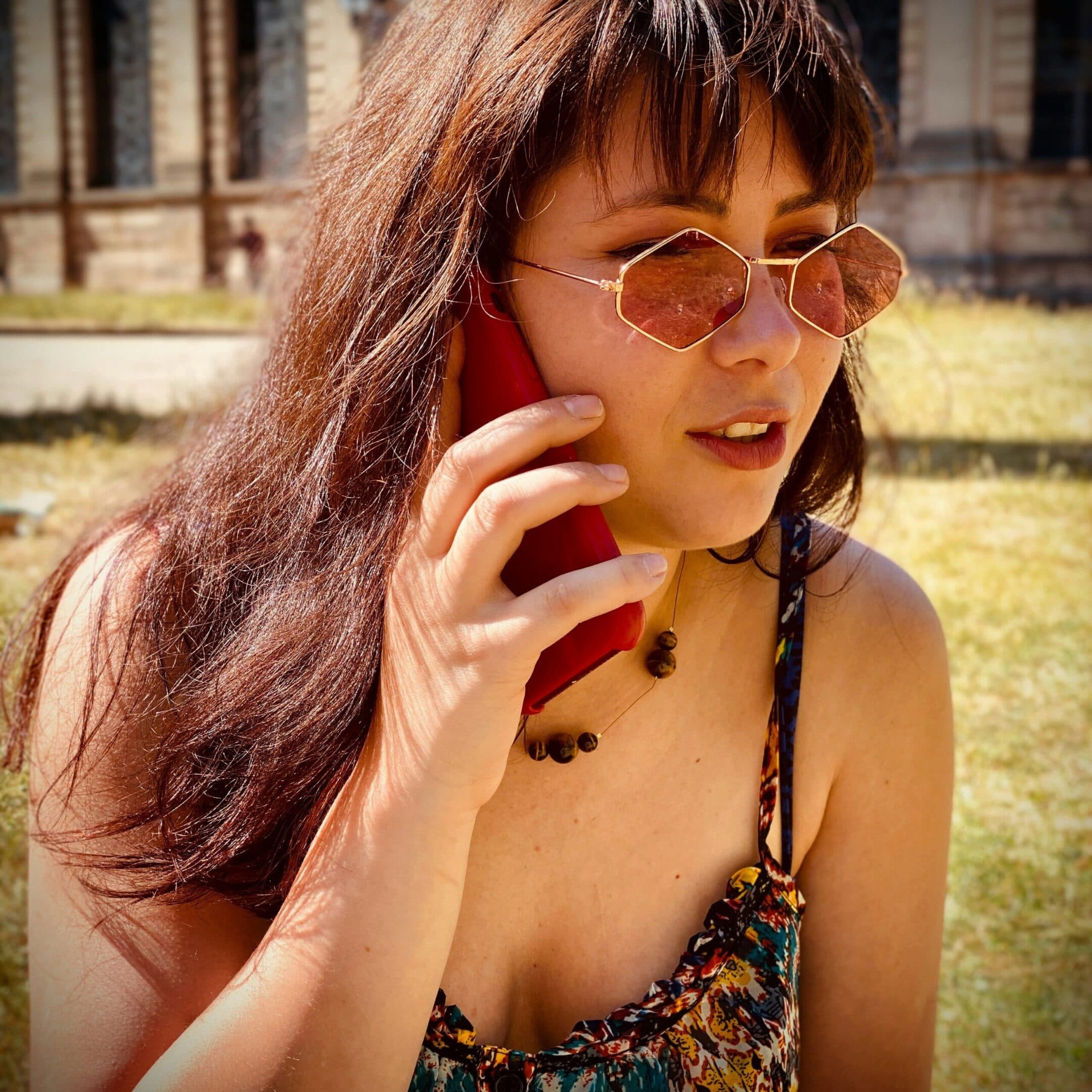 Free Consult A woman with long brown hair and rose-tinted sunglasses is outdoors, holding a red smartphone to her ear. She is wearing a sleeveless floral dress and a beaded necklace, with sunlight highlighting her face.