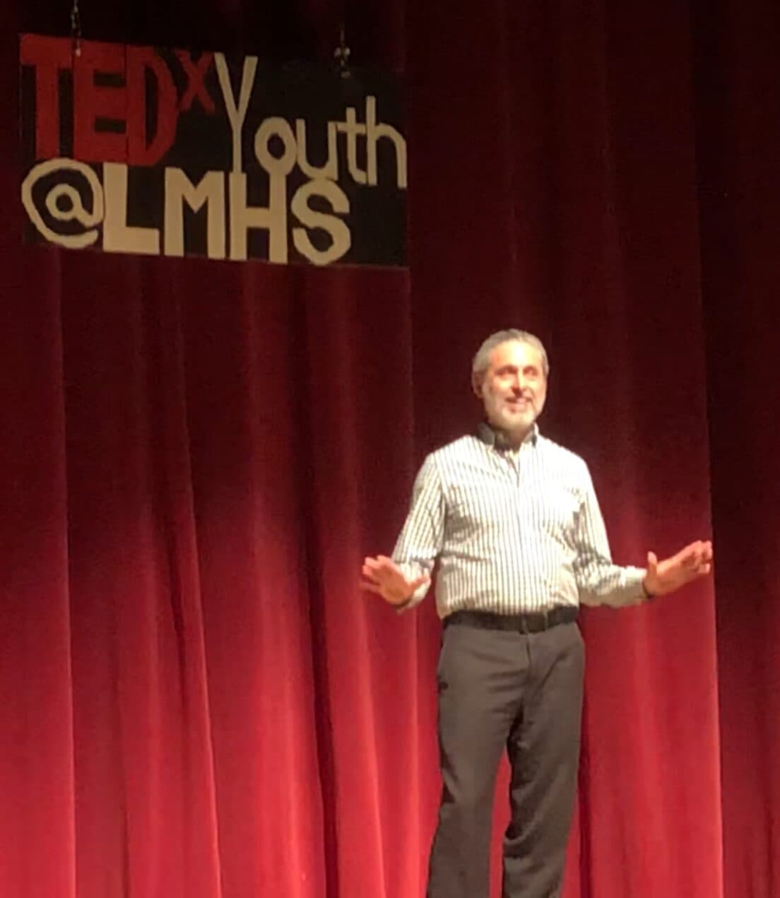 A man stands on stage in front of red curtains, speaking at a TEDxYouth @ LMHS event, with a sign displaying the event name hanging above him.