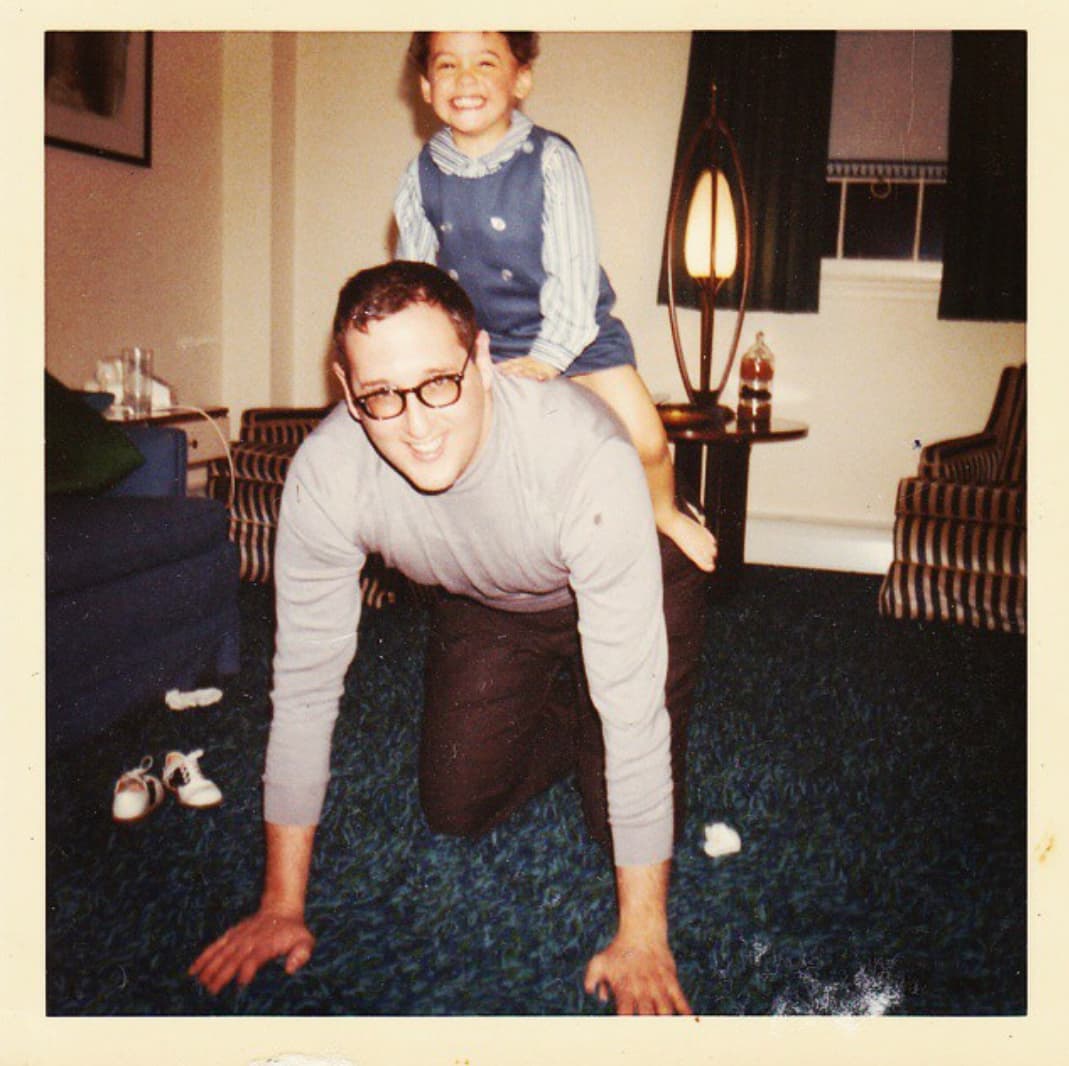 A smiling man with glasses is on all fours on a blue carpet, giving a piggyback ride to a laughing young girl in a blue dress. The cozy room has mid-century furniture and a lamp in the background.